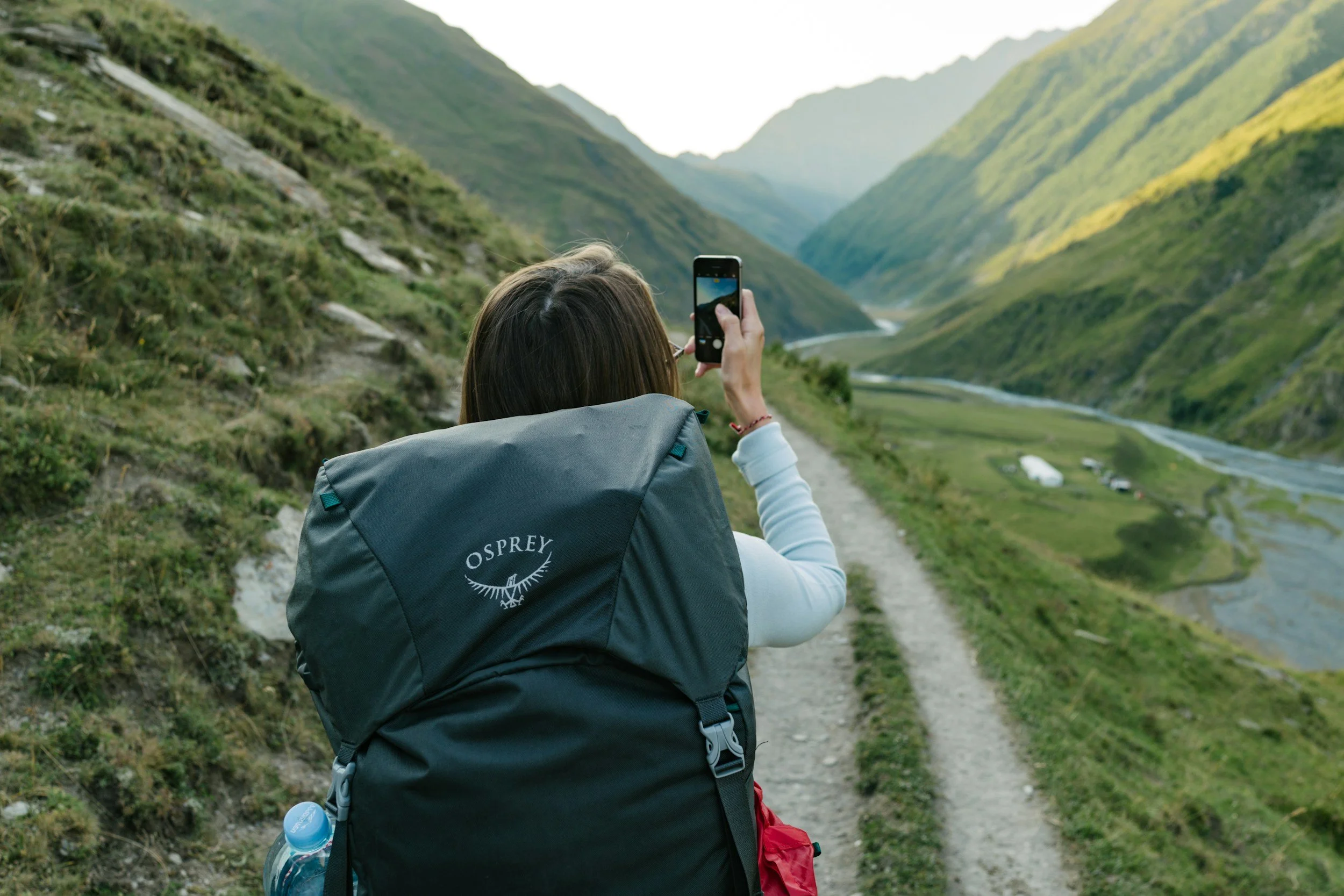 Woman with a backpack taking a photo of a lush green valley with mountains and a river.