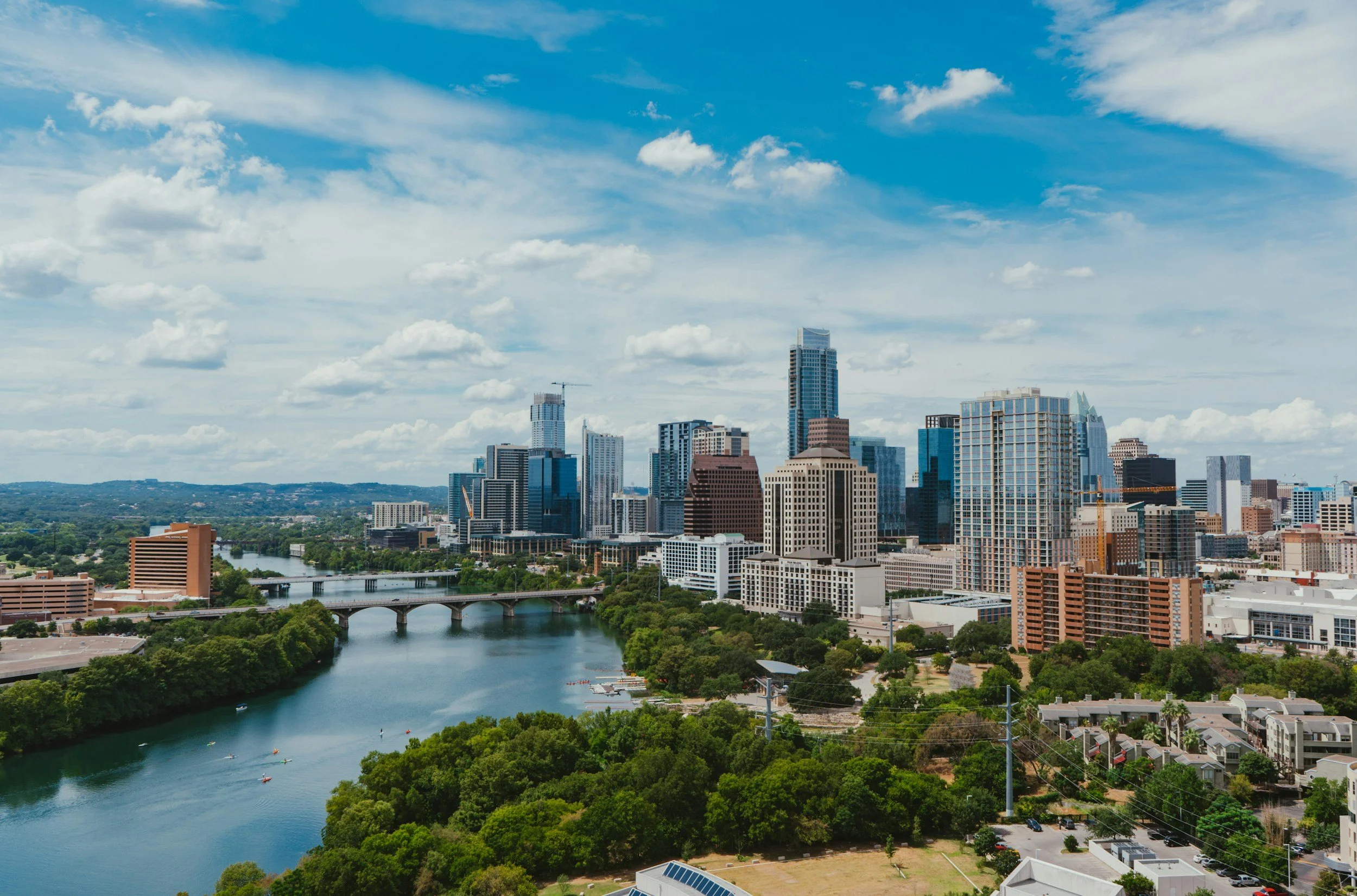 A city skyline with high-rise buildings and skyscrapers, a river with boats, bridges crossing the river, greenery, and a blue sky with clouds.