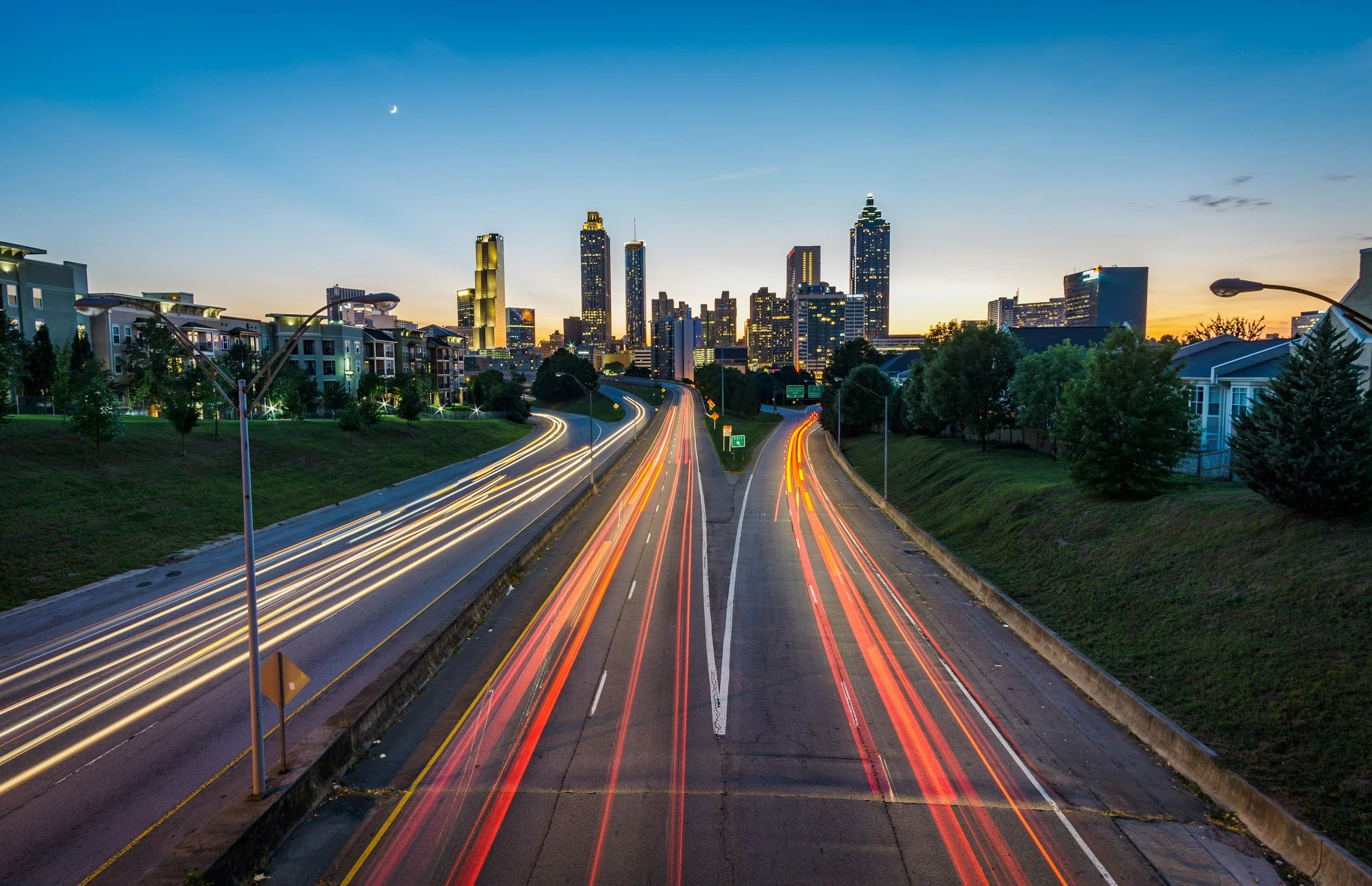 Light trails from moving vehicles on a highway leading into a city skyline at dusk with high-rise buildings and a crescent moon in the sky.