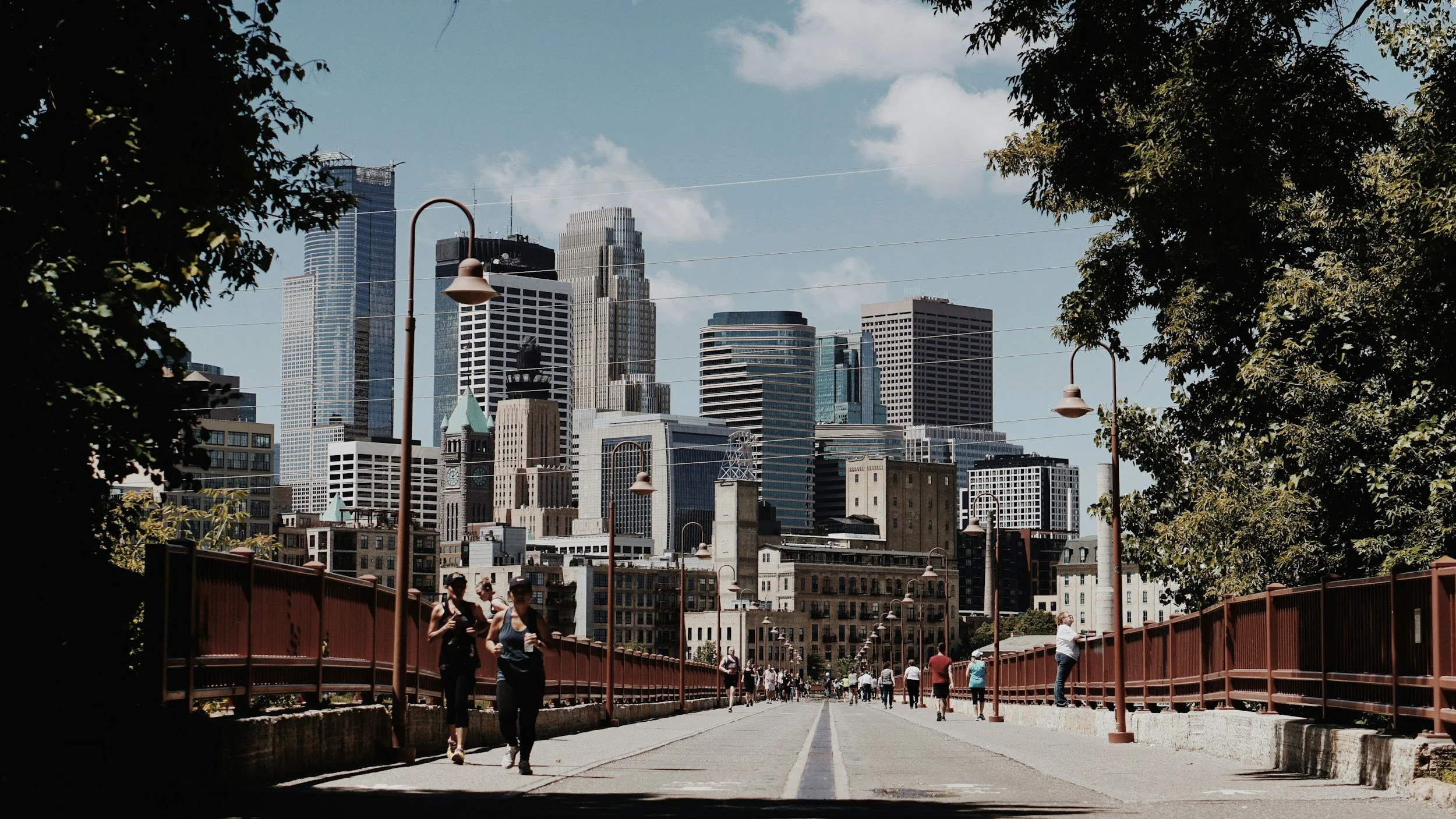 People walking along a pedestrian bridge with a city skyline in the background, featuring tall skyscrapers and historic buildings, with trees on the sides and lampposts lining the bridge.
