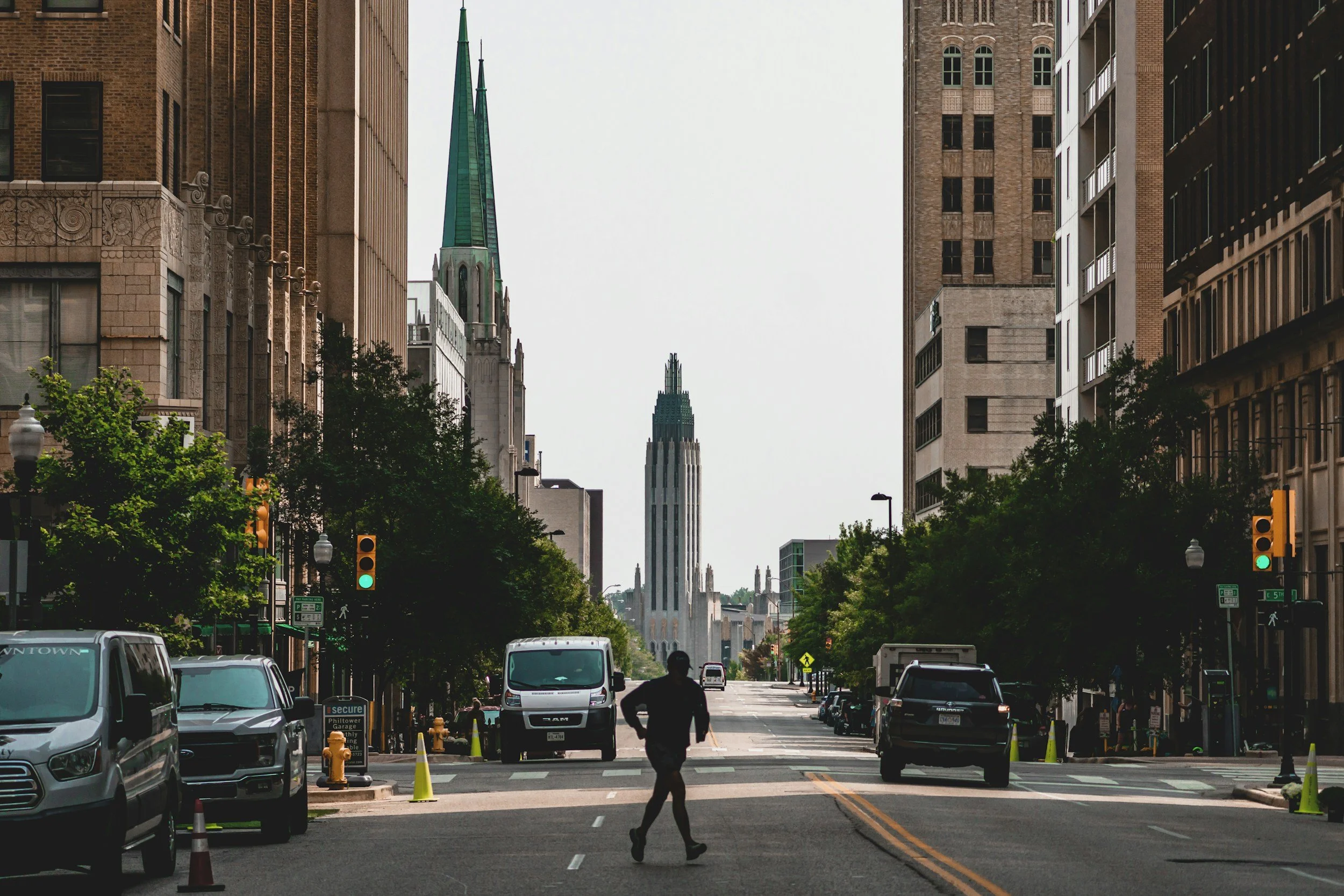 A city street scene with cars parked on both sides, traffic lights, and a person crossing the road. Tall buildings line the street, and a church steeple is visible in the background against a clear sky.