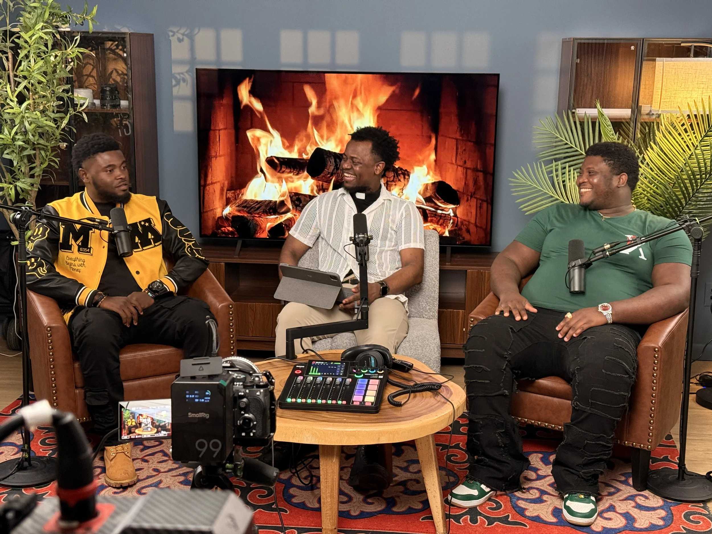 Three men sitting in a recording studio near a table with audio equipment, microphones, and a colorful controller. Behind them is a large screen displaying a fireplace with flames, and there are plants on either side of the room.