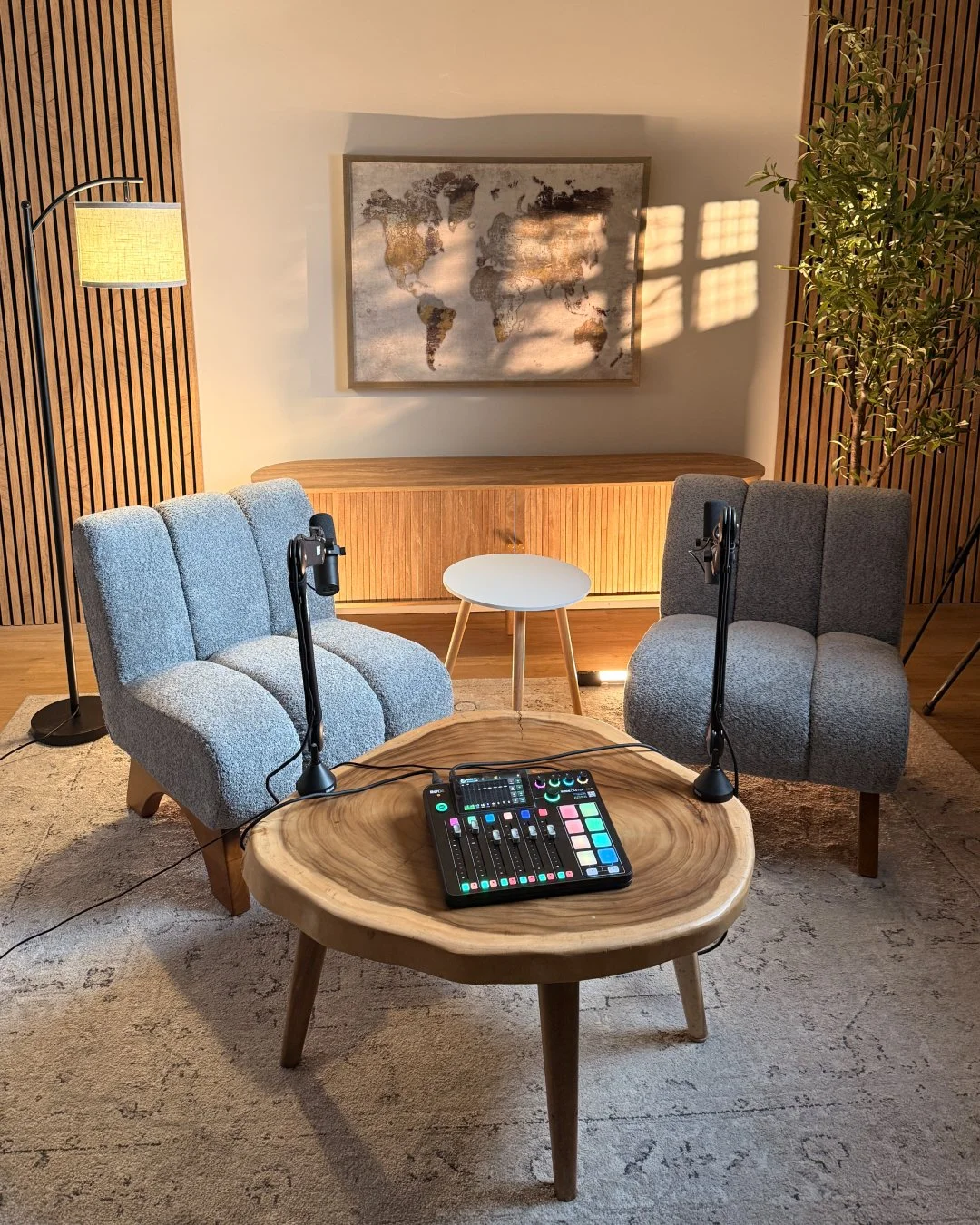 A cozy home studio setup with two grey upholstered chairs facing each other, a small round white side table between them, a wooden table with music or audio equipment, and a large world map artwork on the wall. There are wooden wall panels, a floor lamp, and a tall plant adding warmth to the room.