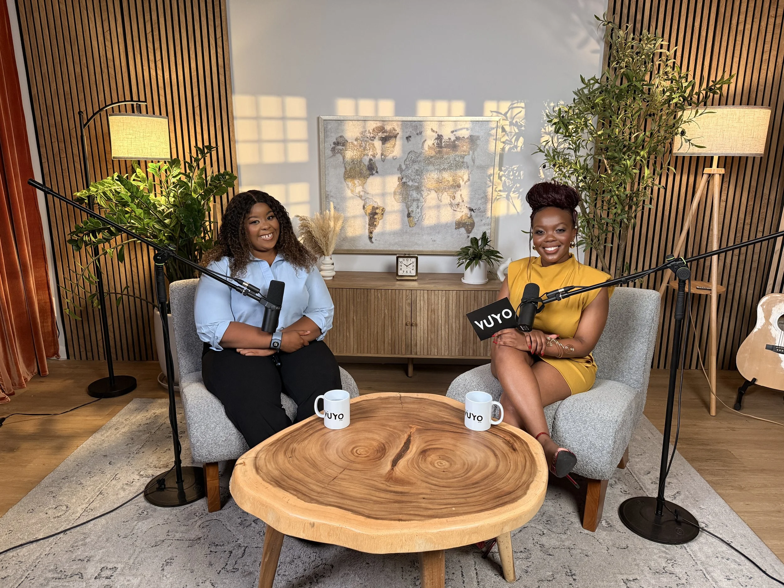 Two women sitting on a cozy grey sofa with a round wooden coffee table in front of them, microphones, coffee mugs labeled 'Vuyo,' and a casual background with wall art, plants, lamps, and wood paneling.