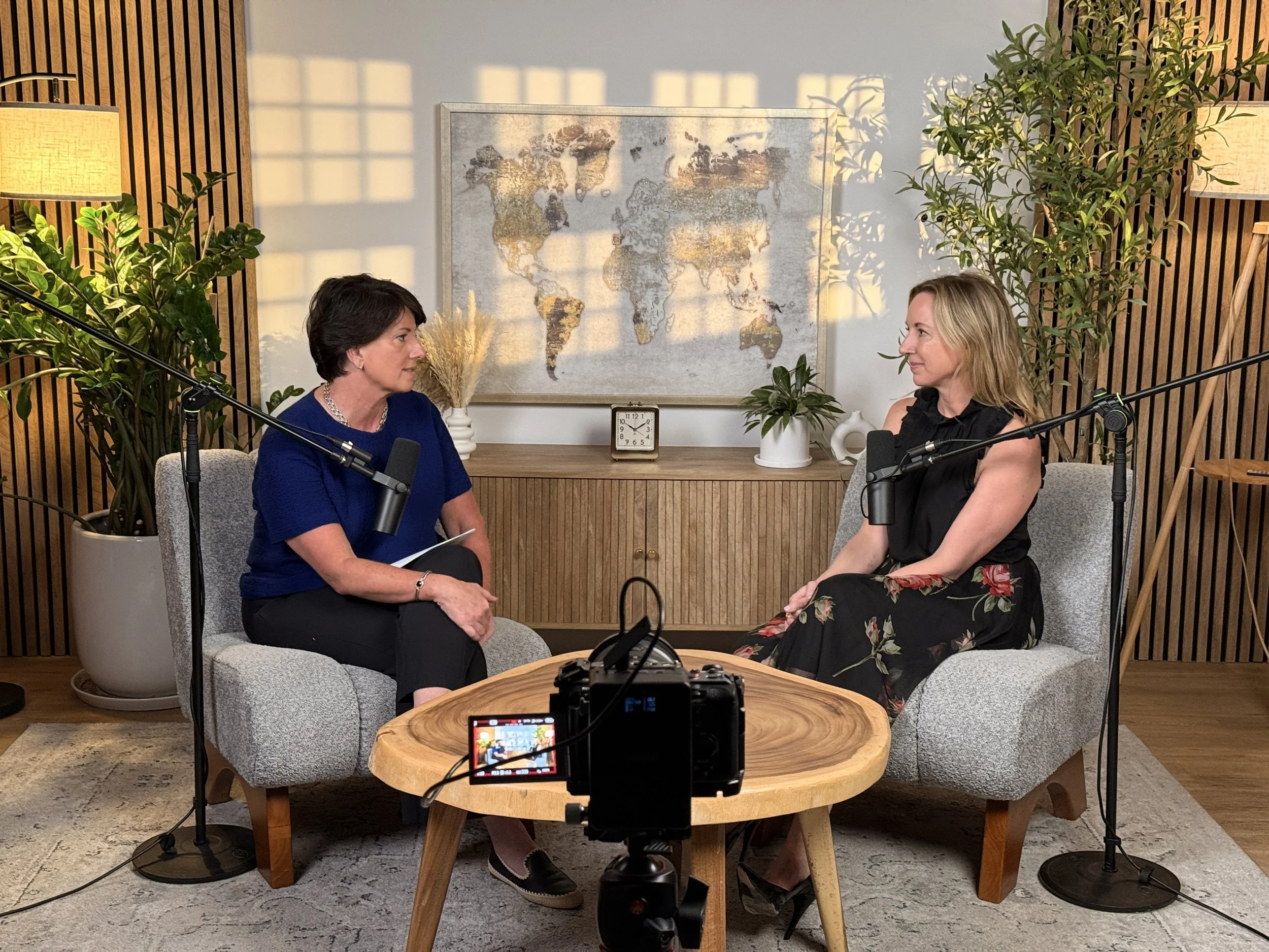 Two women sit facing each other in a room decorated with plants and a world map. They are surrounded by recording equipment, including microphones and a camera, indicating they are recording or podcasting.