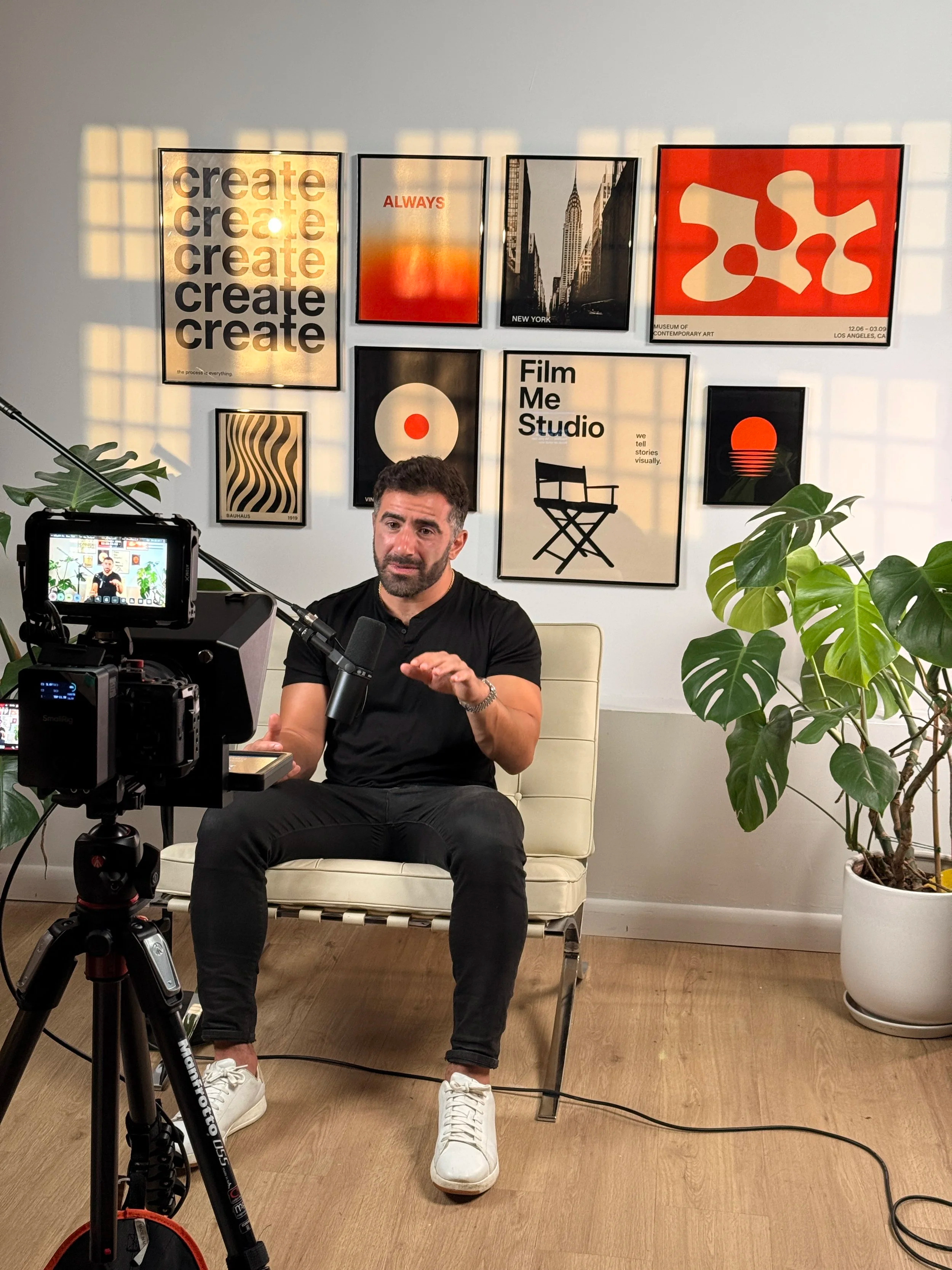 A man sitting on a cream-colored chair in front of a camera setup, speaking into a microphone, with framed artwork and plants in the background.