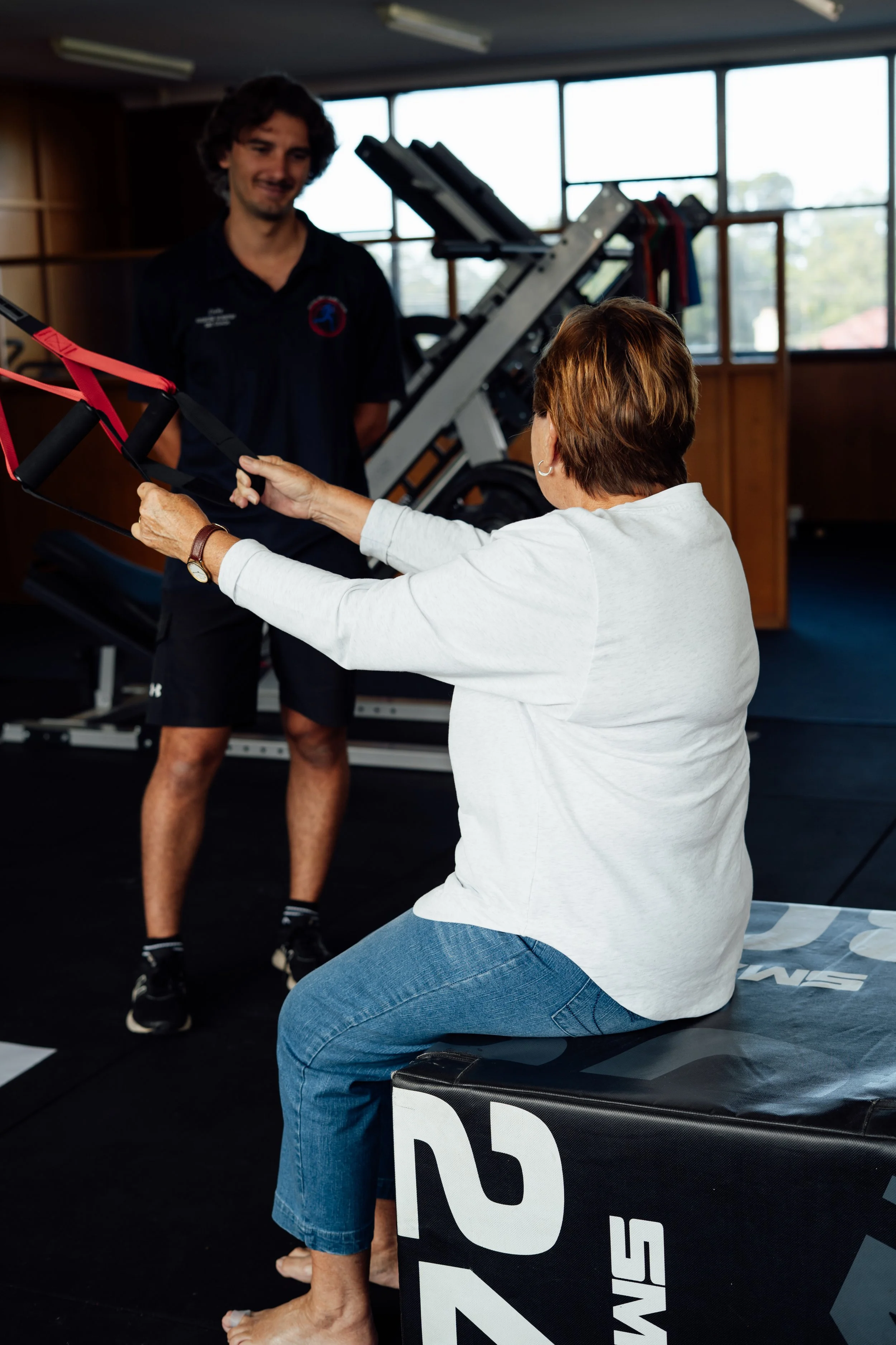 A woman is sitting on a fitness bench while holding resistance bands, looking at a smiling man standing in front of her in a gym with large windows and exercise equipment in the background.
