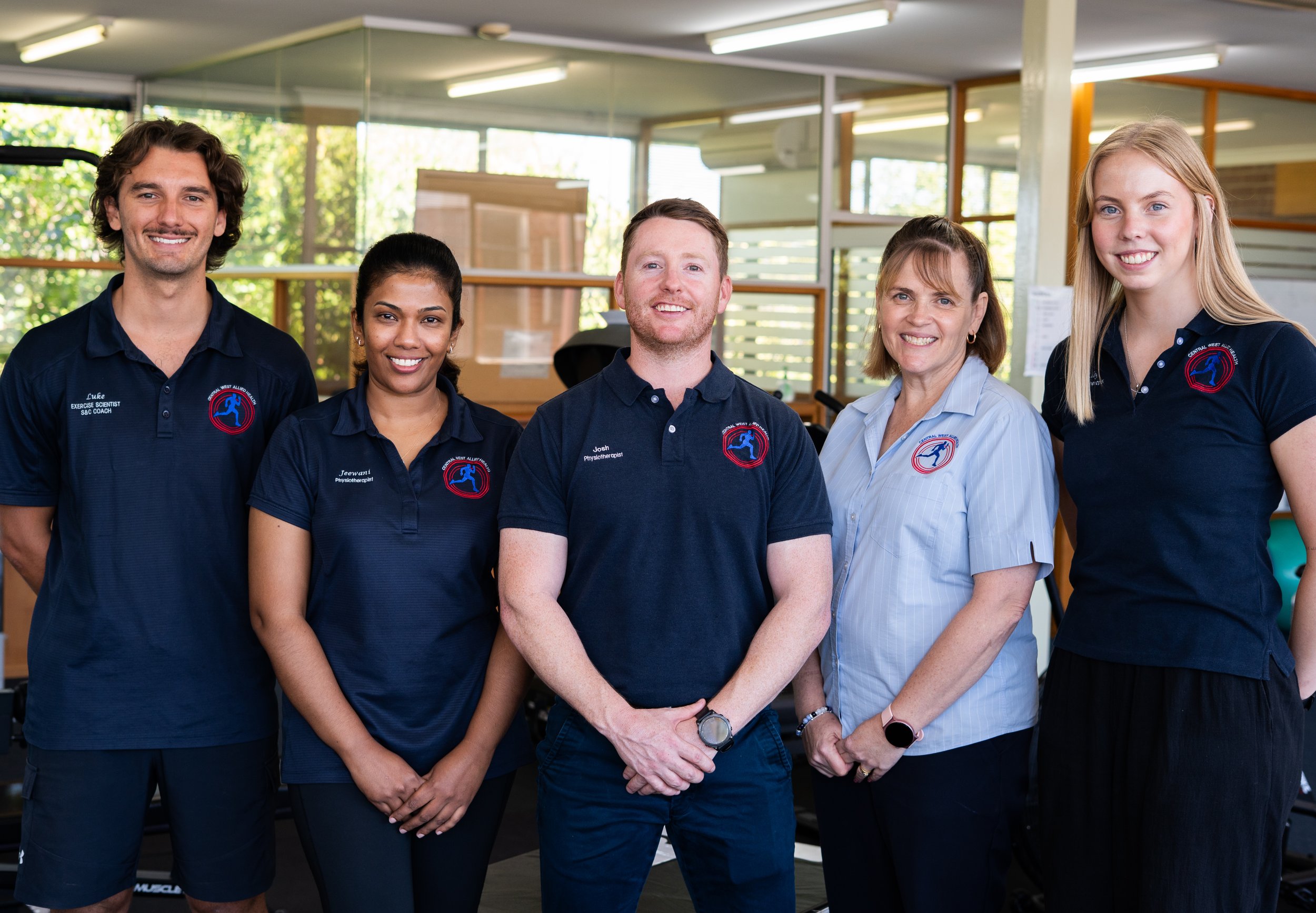 Group of five diverse fitness professionals standing indoors, smiling at the camera, in a gym or fitness center.