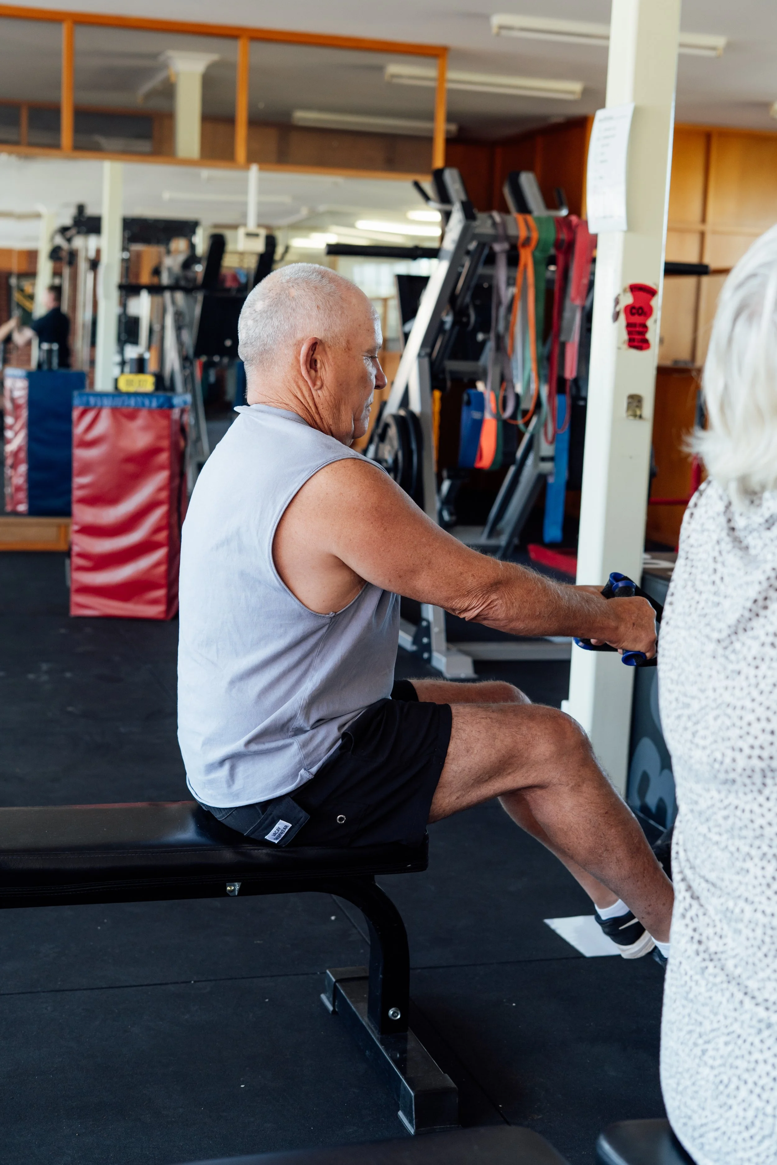 An older man with a shaved head sitting on a workout bench in a gym, engaged in physical activity, with gym equipment and a person with white hair visible nearby.