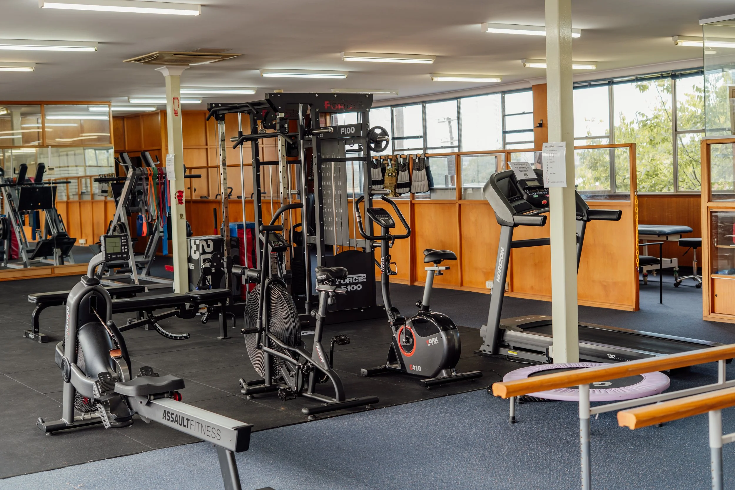 Interior view of a gym with various exercise equipment including stationary bikes, a treadmill, and weightlifting machines, with large windows letting in natural light.