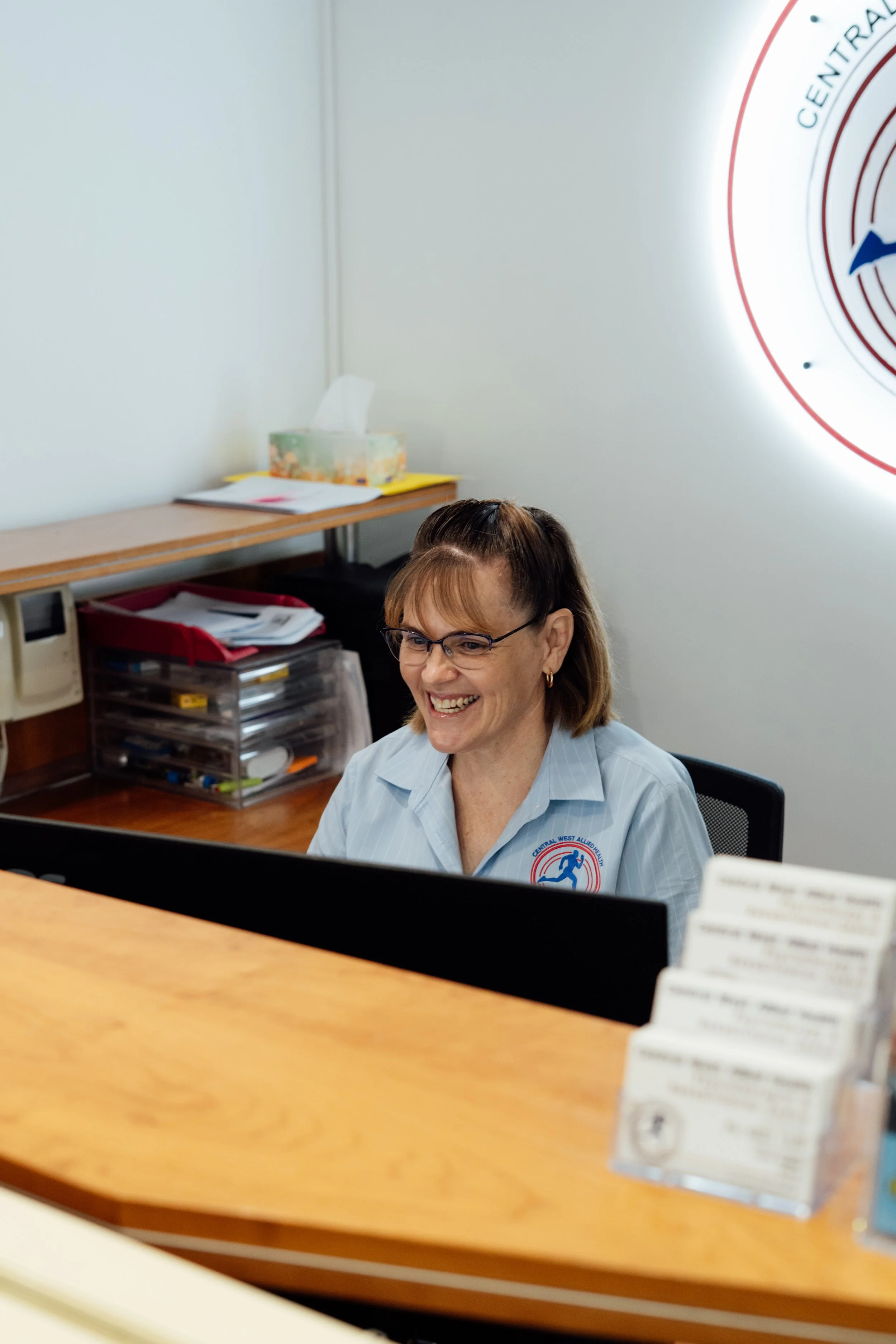 A woman with glasses smiling sitting at a desk in an office, with a tissue box, papers, and office supplies in the background, and a logo on the wall.