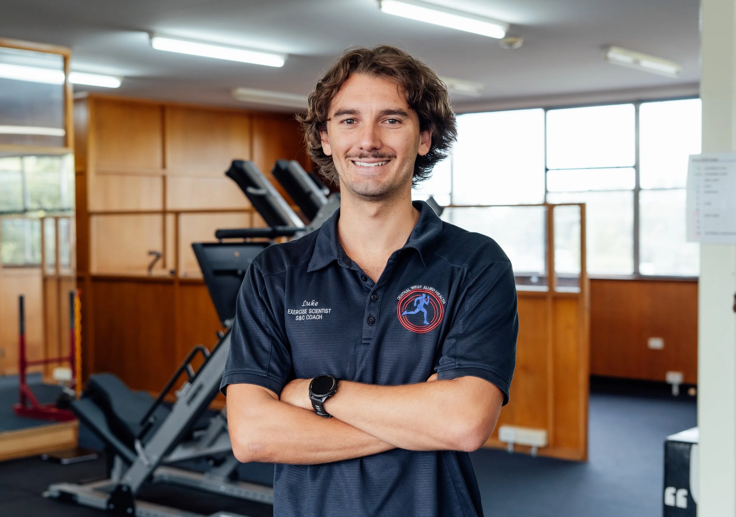 A smiling man with curly brown hair wearing a dark polo shirt with a logo, standing with arms crossed in a gym near exercise equipment.