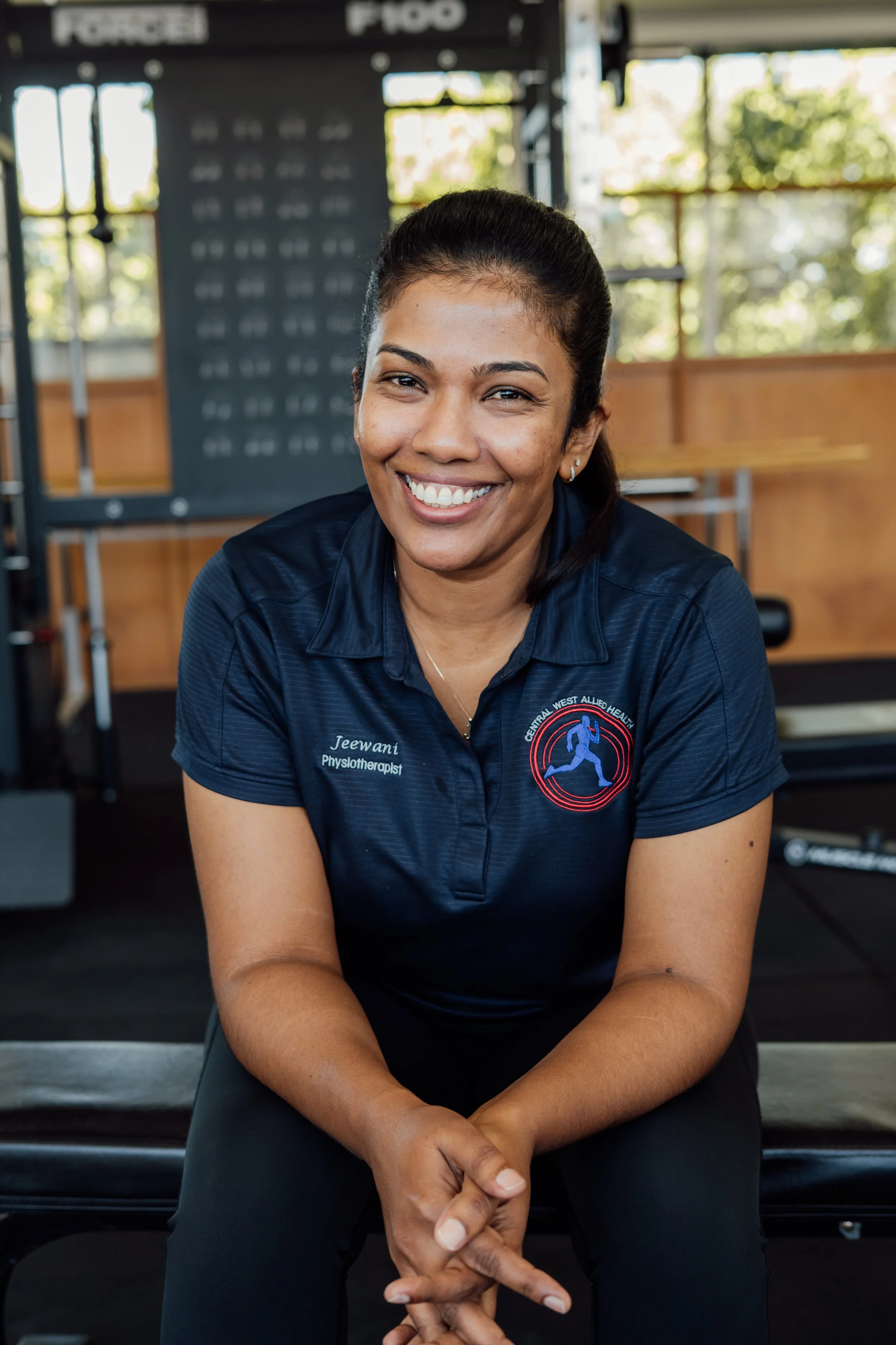 A smiling woman with dark hair pulled back, wearing a navy blue polo shirt with a logo, sitting in a gym with equipment and large windows in the background.