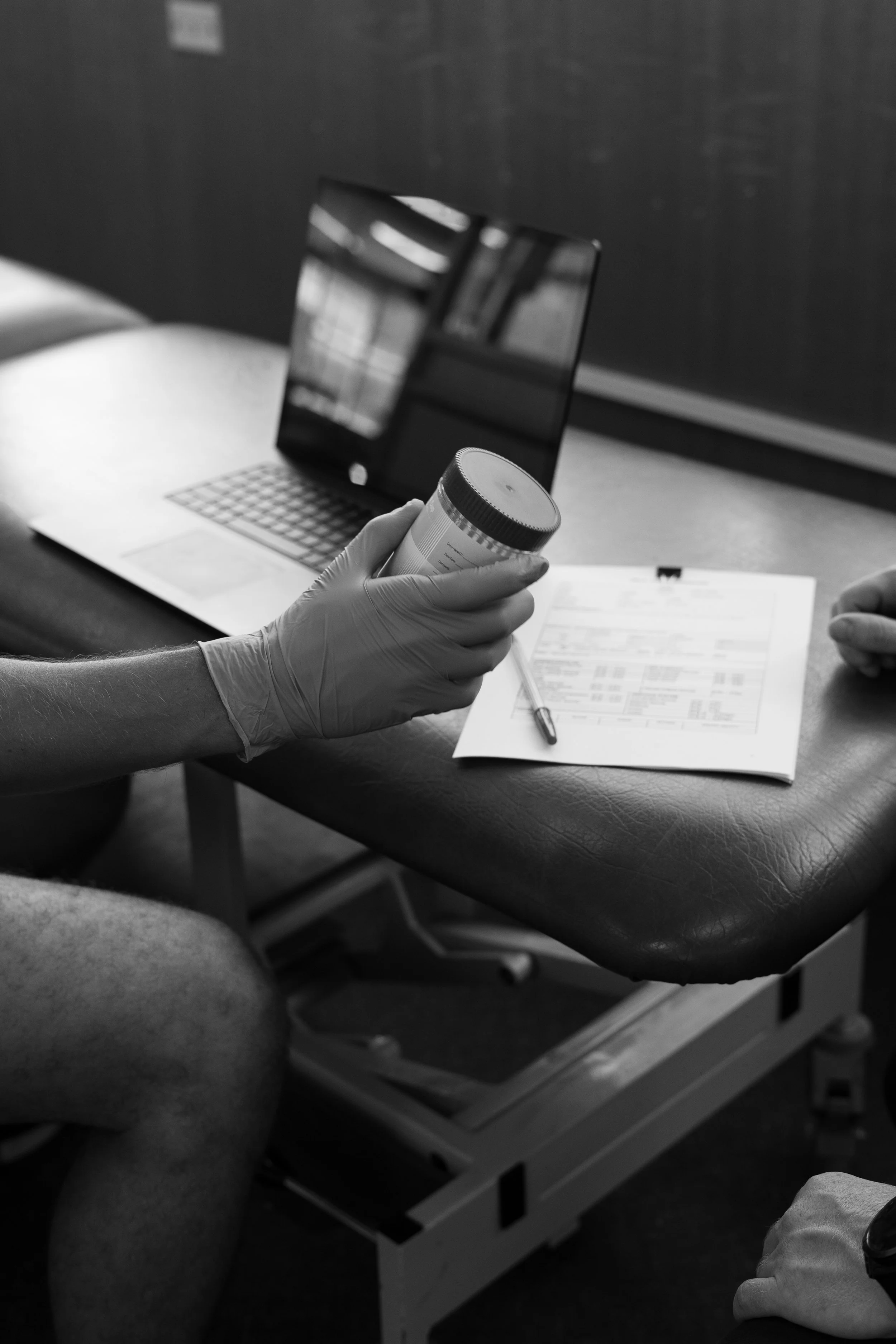 A person wearing gloves holding a prescription pill bottle in front of a laptop, with a paper and pen on a table.