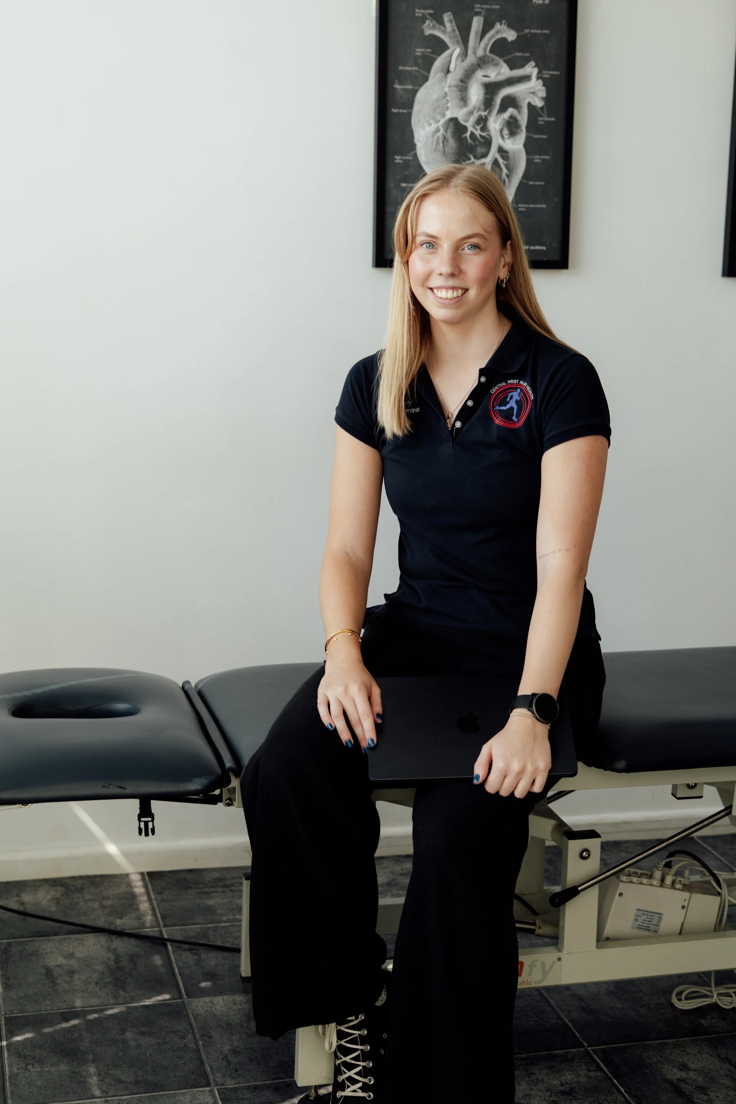 A young woman with long blonde hair, wearing a black polo shirt with a logo and text, black pants, white lace-up boots, a watch, earrings, and a bracelet, sitting on a medical examination table in a clinic. She is smiling and holding a silver laptop on her lap. There is a diagram of a human heart on the wall behind her. The setting appears to be a healthcare or physical therapy clinic.