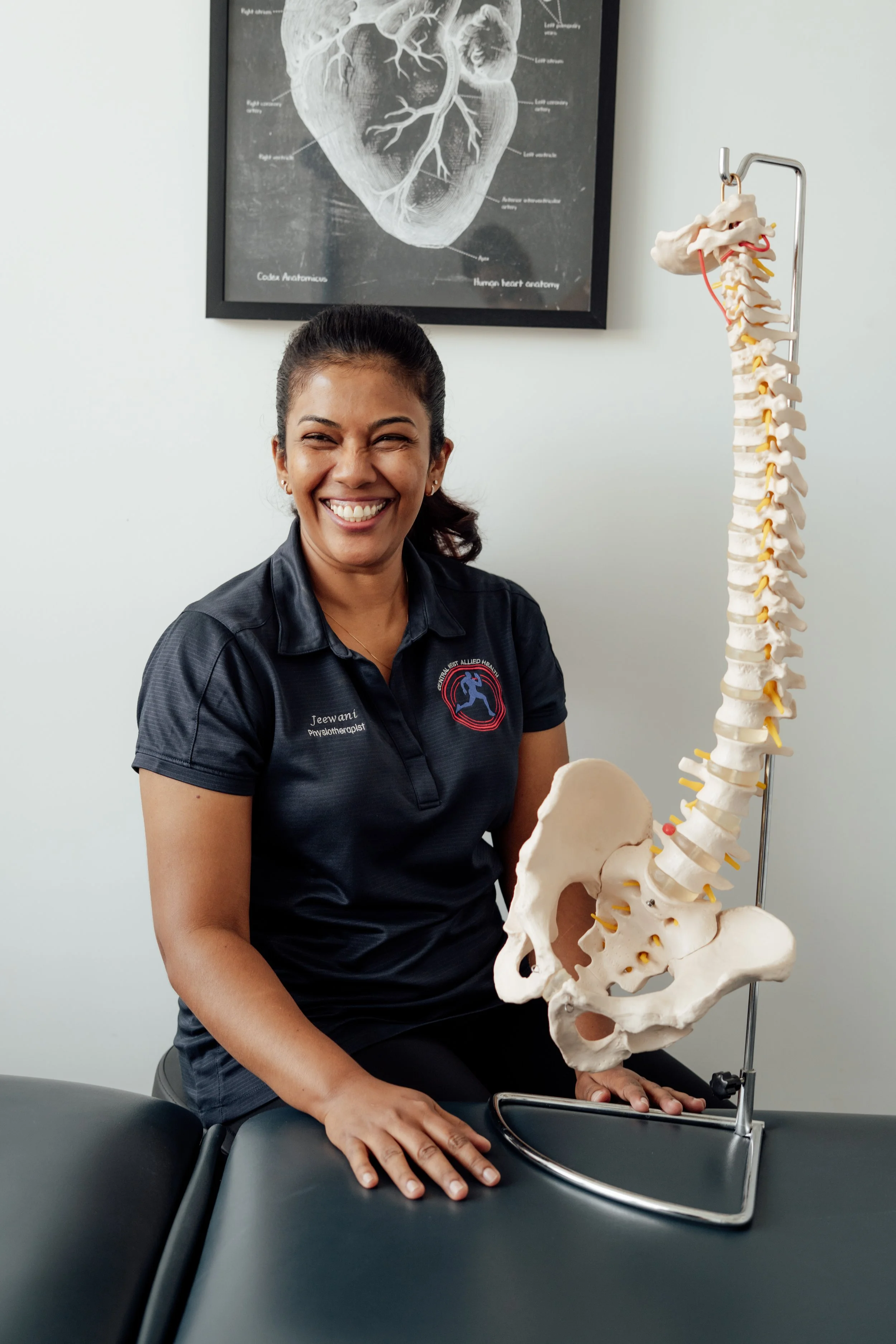A woman smiling sitting beside a model of a human spine and pelvis in a medical office or classroom setting.
