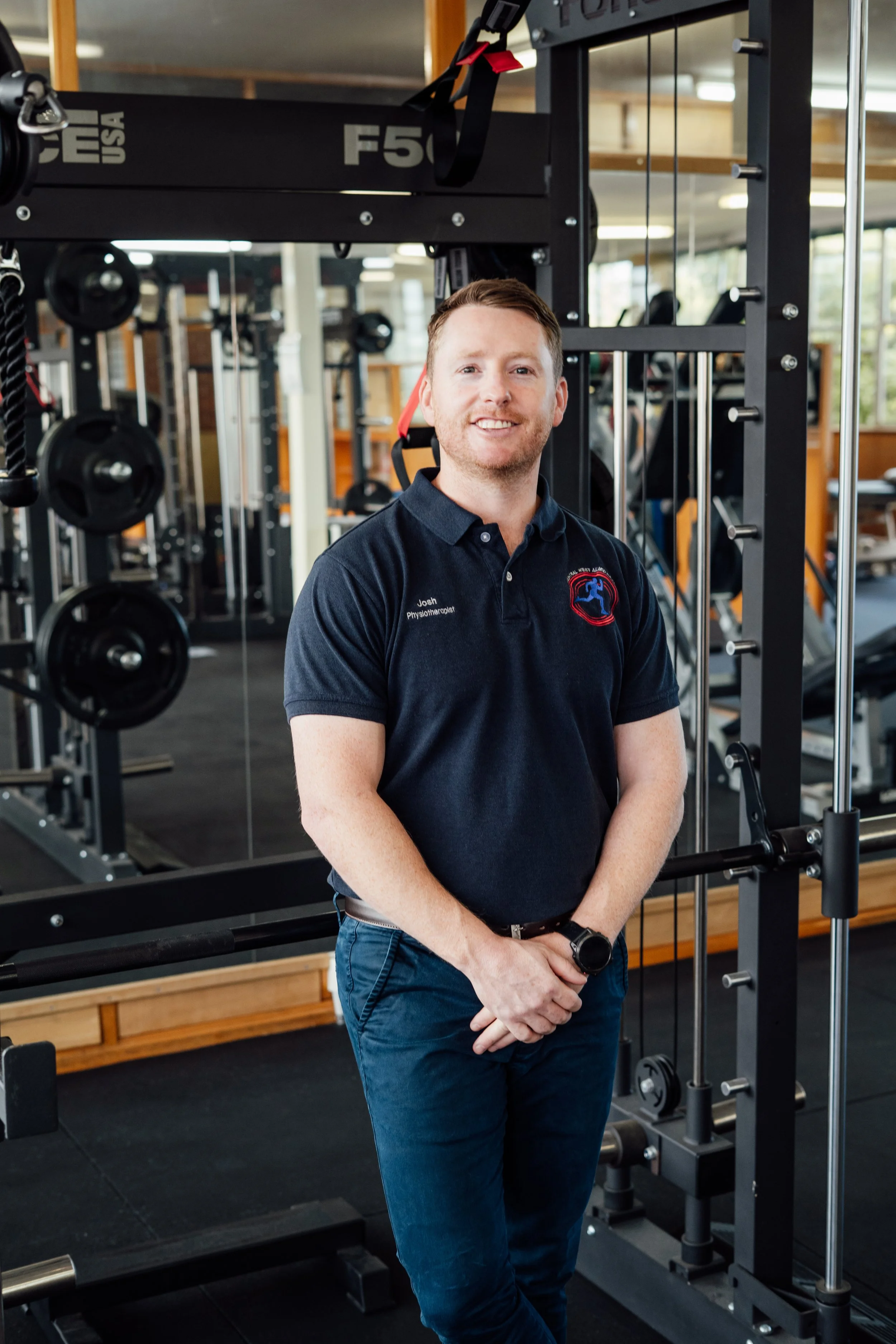 A man with short brown hair and a beard, wearing a navy blue polo shirt with a logo and text that says "Josh Physical Therapist," standing in a gym near fitness equipment with weight racks and machinery, smiling at the camera.