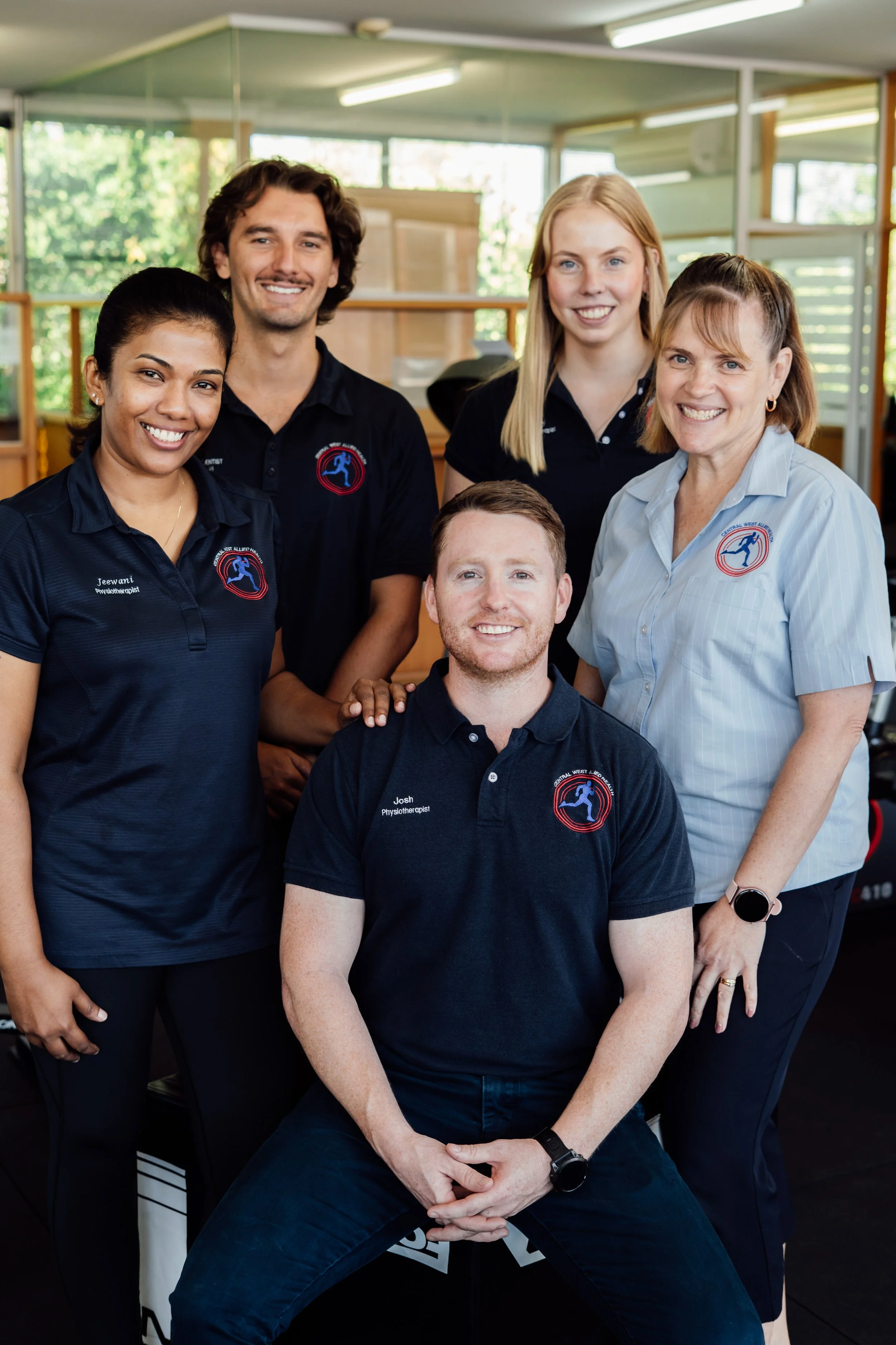 A group of five people, four women and one man, are smiling and posing together in an indoor setting with large windows. Three people are standing behind a seated man, with each person wearing dark shirts with a logo on them. The woman on the right wears a light-colored shirt with a logo, and the woman on the left has a name tag that reads 'Jeewani' and 'Physiotherapist'. The man in front is looking directly at the camera with a smile, while the others are standing behind him, also smiling.