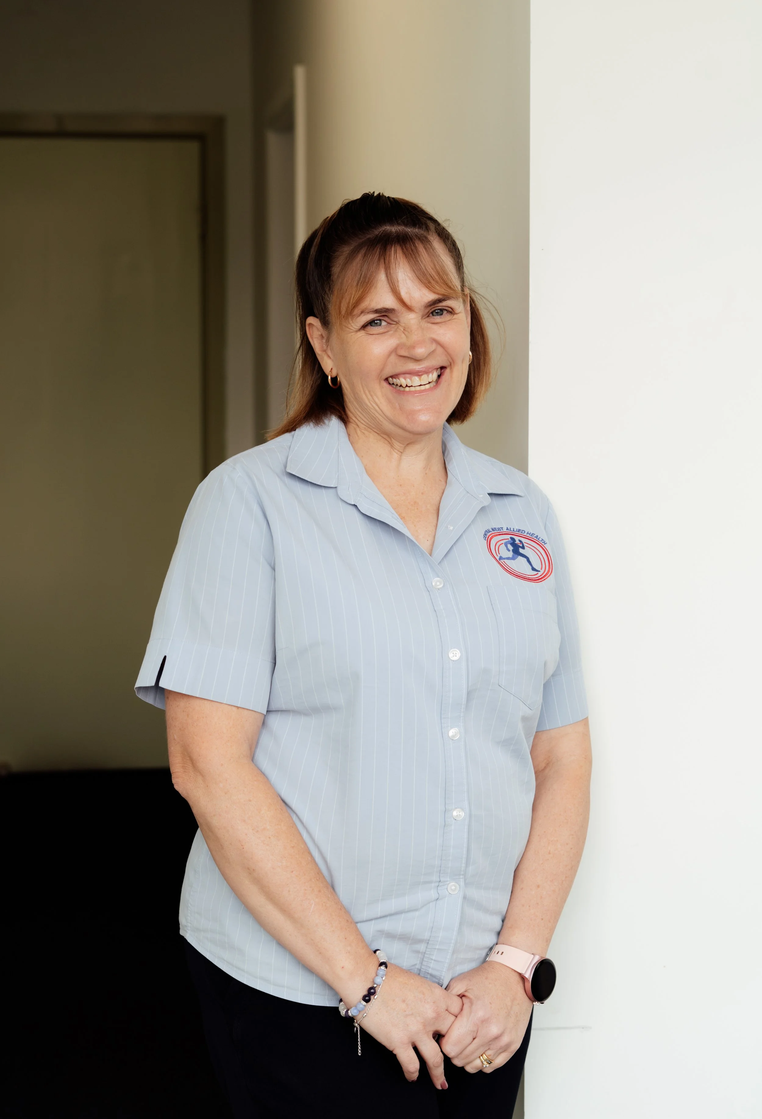 A smiling woman in a light blue striped shirt with a logo, standing against a plain wall, wearing jewelry and a smartwatch.