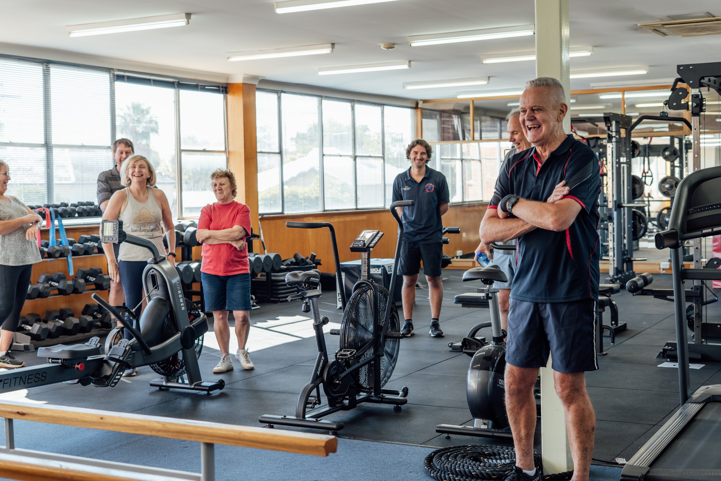 Group of older adults and trainers in a gym, smiling and talking, with exercise equipment around.