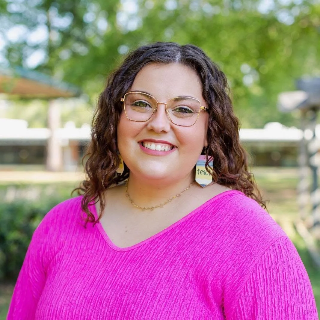 A woman with curly brown hair wearing glasses and earrings smiling outdoors in a park with green trees in the background, dressed in a bright pink top.