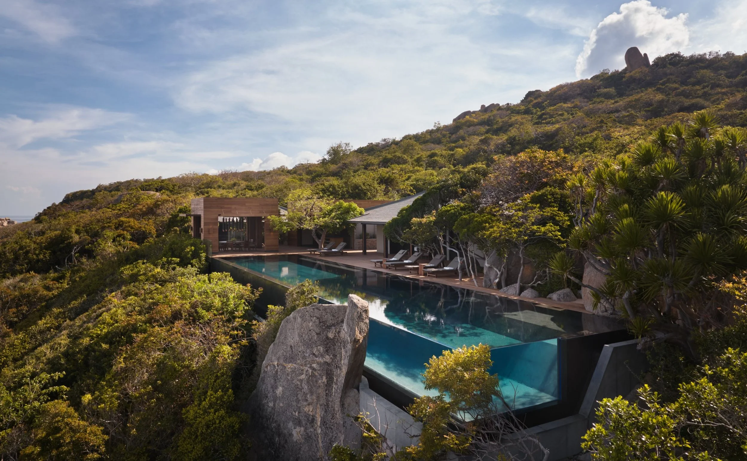 Luxury modern house with an infinity pool surrounded by lush trees and hillside with rocks.