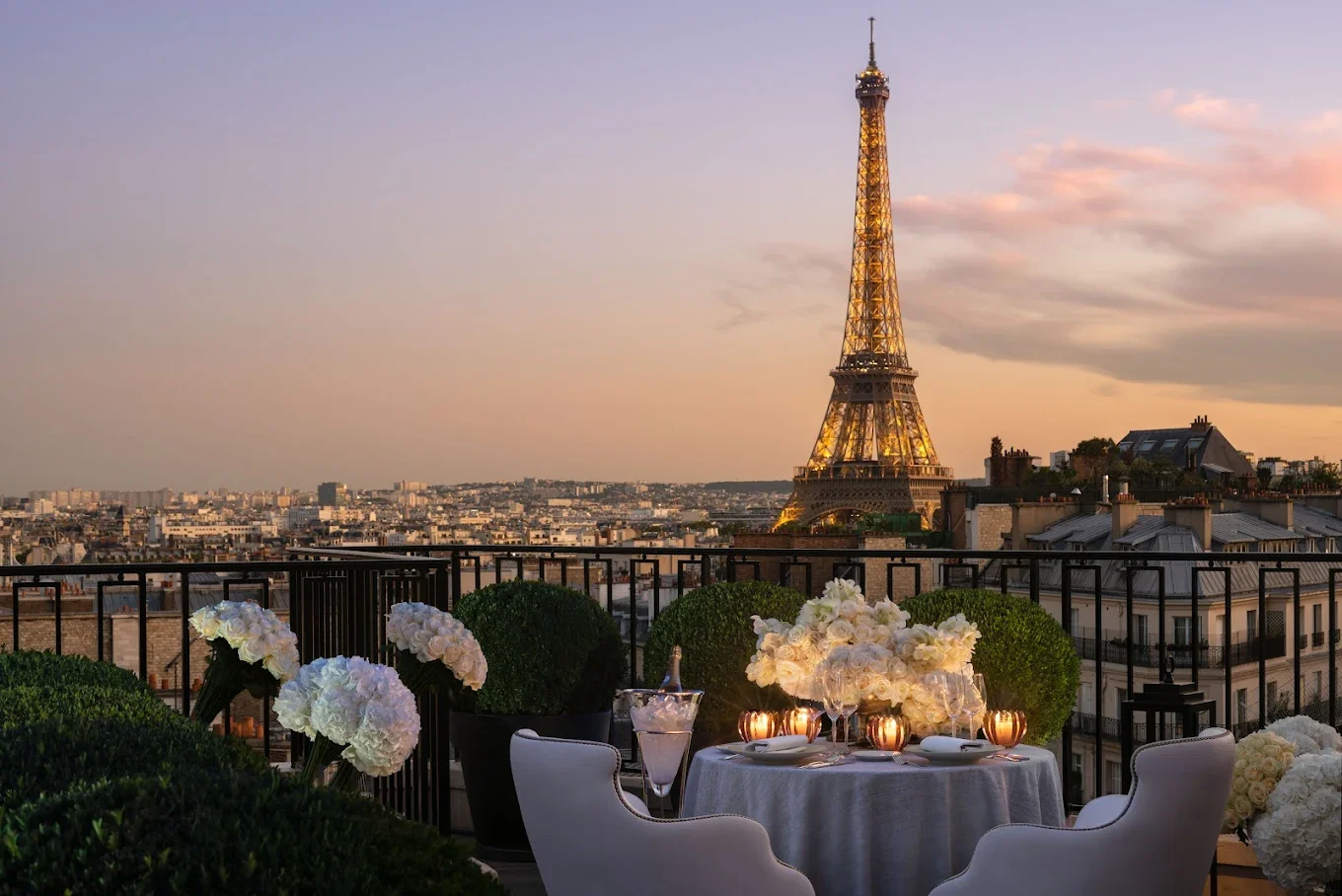 Rooftop dining setup with a table and chairs, decorated with white flowers and surrounded by potted plants, overlooking the illuminated Eiffel Tower during sunset in Paris.
