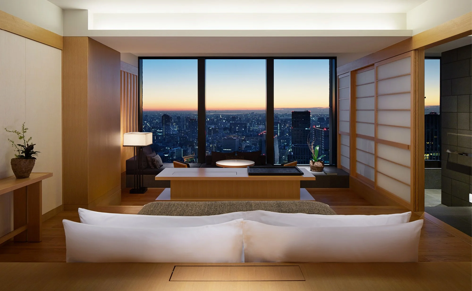 Interior view of a modern high-rise apartment living room during sunset with large windows showing a city skyline.