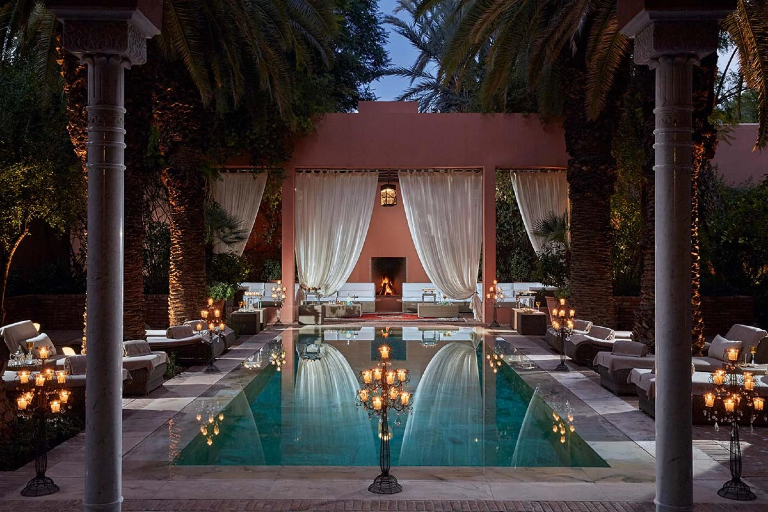 A luxurious outdoor pool area at dusk, with the pool reflecting string lights and chandeliers, surrounded by lounge chairs, and a backdrop of a pink building with white curtains and a fireplace.