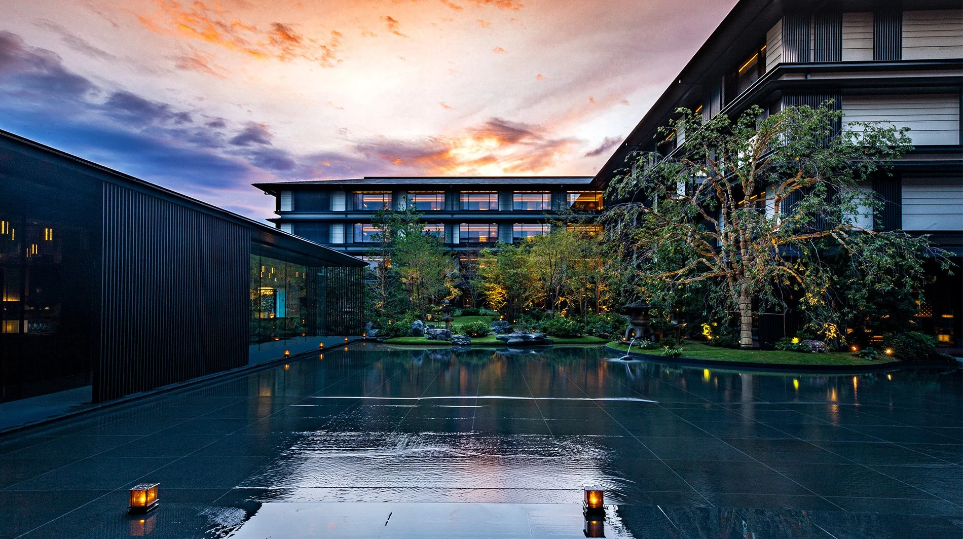 Modern building courtyard with reflective wet black tile ground, illuminated garden, trees, and sunset sky.