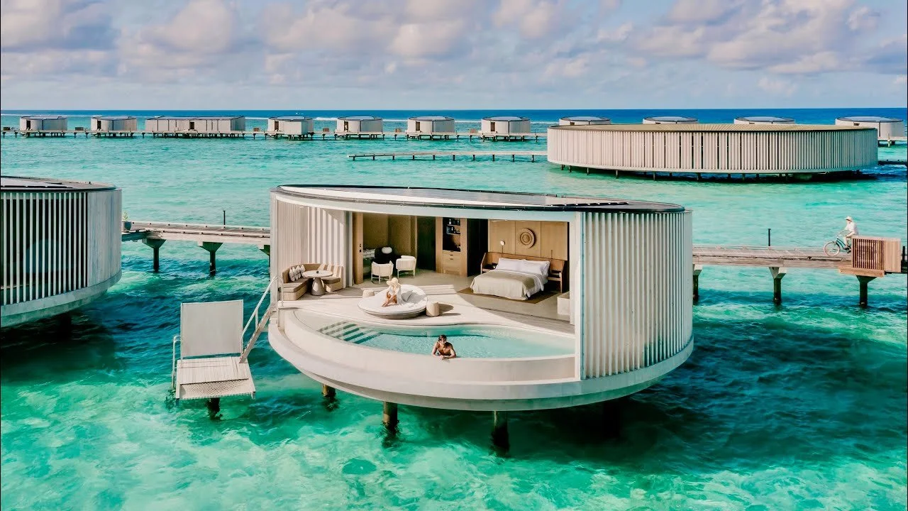 Overwater bungalow with a circular design and a small pool, situated on turquoise ocean water, with a sky filled with clouds in the background.