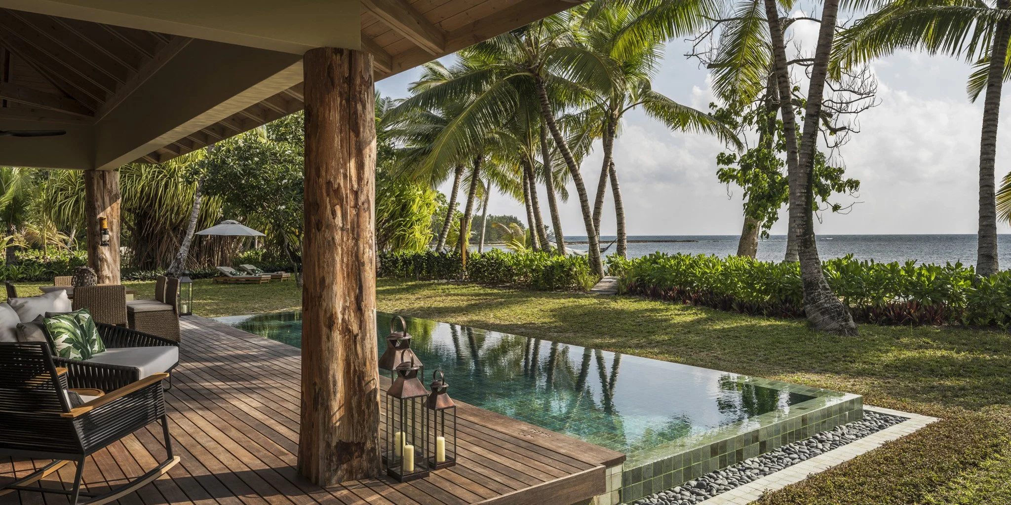 A tropical outdoor scene with a wooden deck, a small pool, and lush greenery including palm trees, with the ocean in the background.