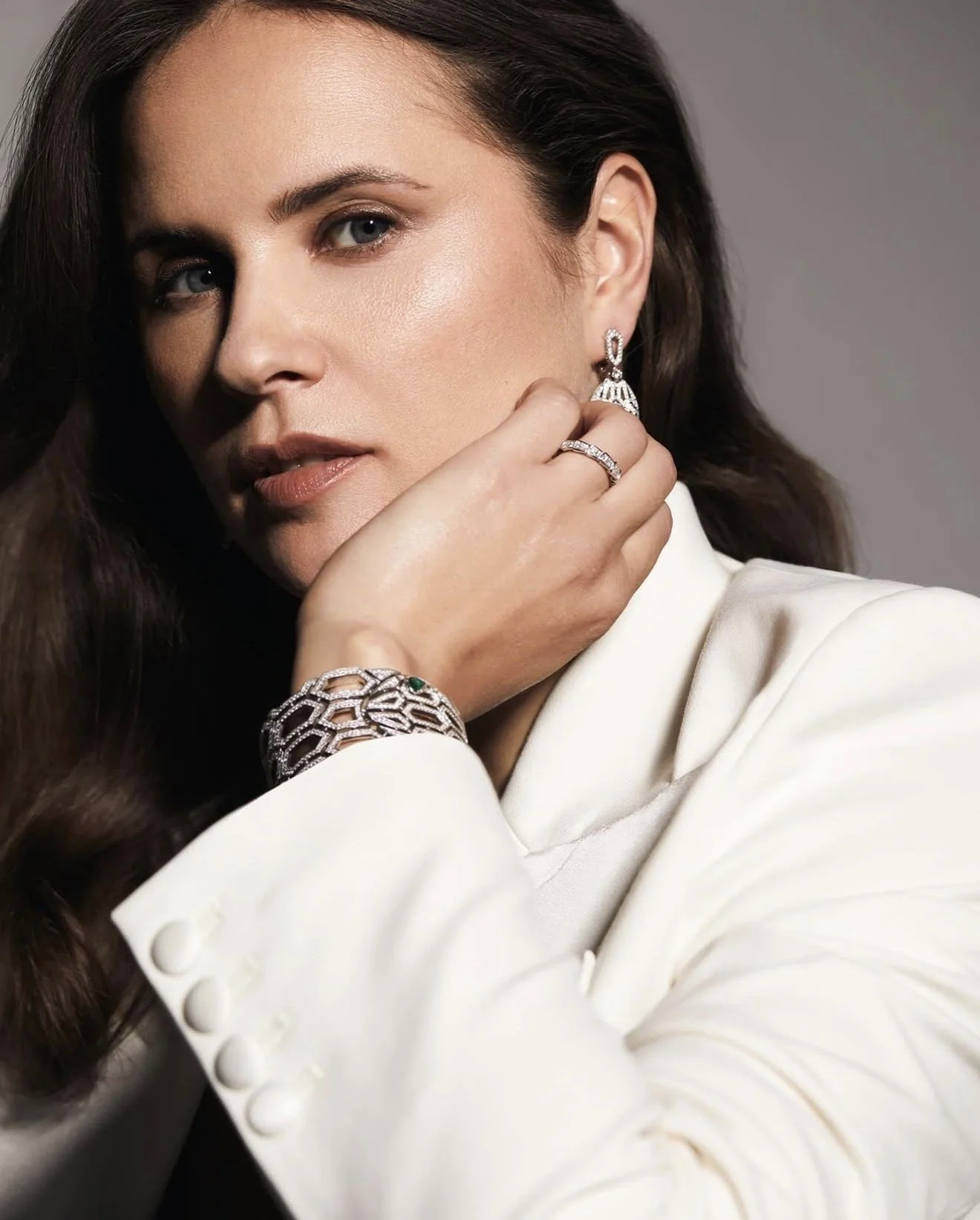 A woman with brown hair and blue eyes wearing elegant jewelry, including earrings, a bracelet, and a ring, dressed in a white blazer, with a neutral gray background.