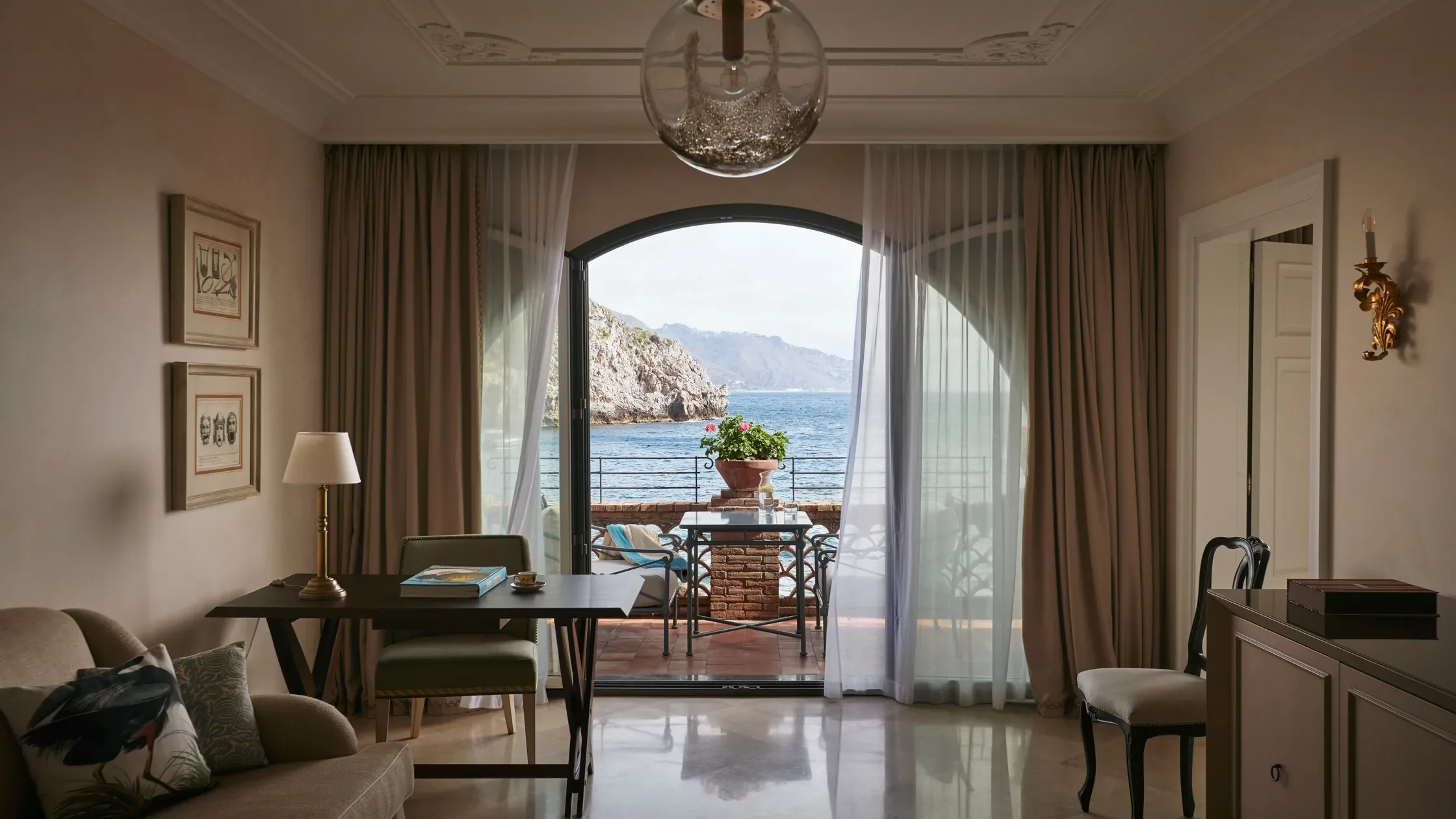 Room with a large arched window opening to a balcony overlooking the sea, with a view of rocky cliffs and mountains in the distance. Inside, there is a beige sofa with decorative pillows, a wooden table with books and a lamp, and another chair near a cabinet. The balcony has a table with a potted plant and two chairs.