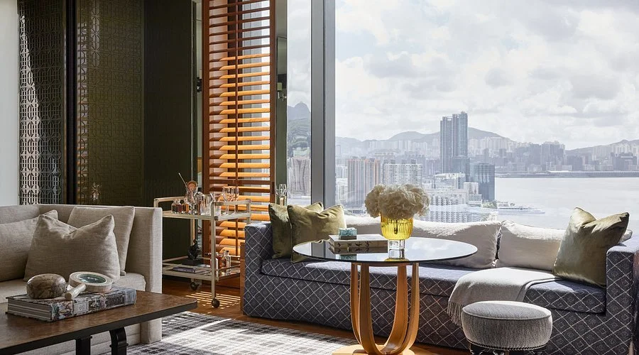 Living room with large windows overlooking a cityscape and harbor, decorated with a beige sofa, blue patterned sectional, round coffee table with flowers, and bar cart with glasses.