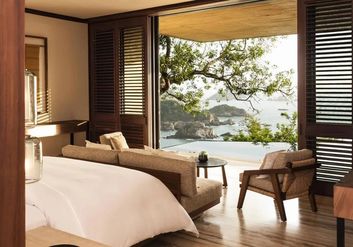 A hotel room with a large bed, beige sofa, and armchair, facing an open balcony with a view of trees, rocky shoreline, and water under a wooden ceiling.