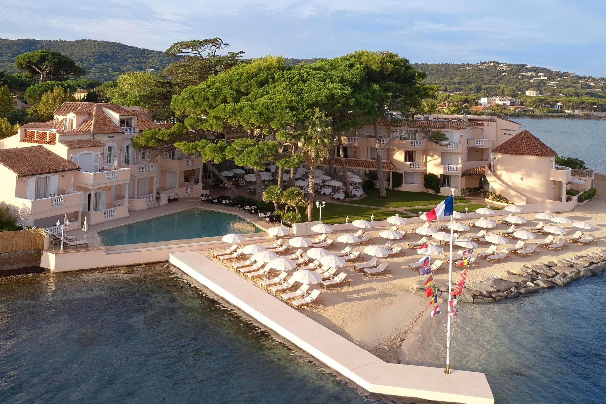 Aerial view of a luxury seaside resort with a swimming pool, numerous umbrellas and lounge chairs on a sandy beach, lush green trees, and a calm body of water in the background, along with a flagpole with multiple national flags.
