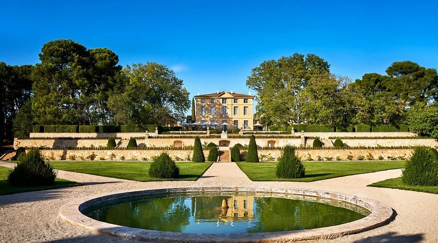 A large historic mansion surrounded by landscaped gardens, trees, and a pond in the foreground on a bright, clear day.
