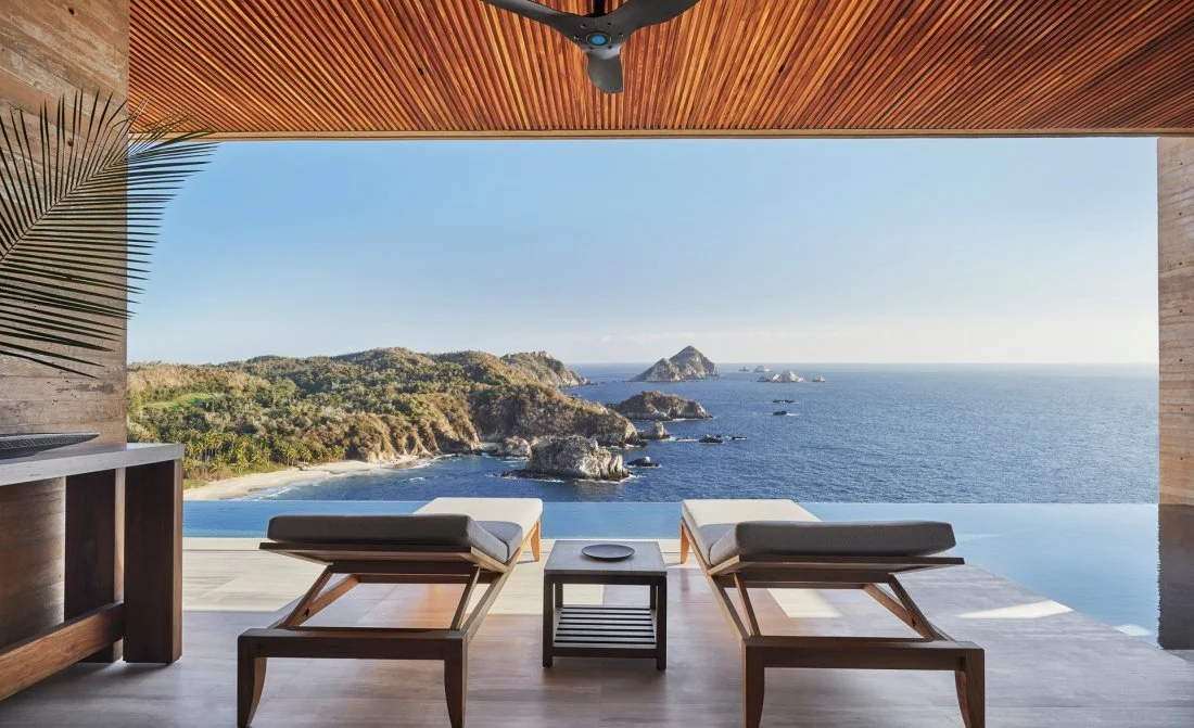 A luxurious outdoor deck with two lounge chairs and a small table, overlooking a scenic ocean view of rocky islands and a clear blue sky.