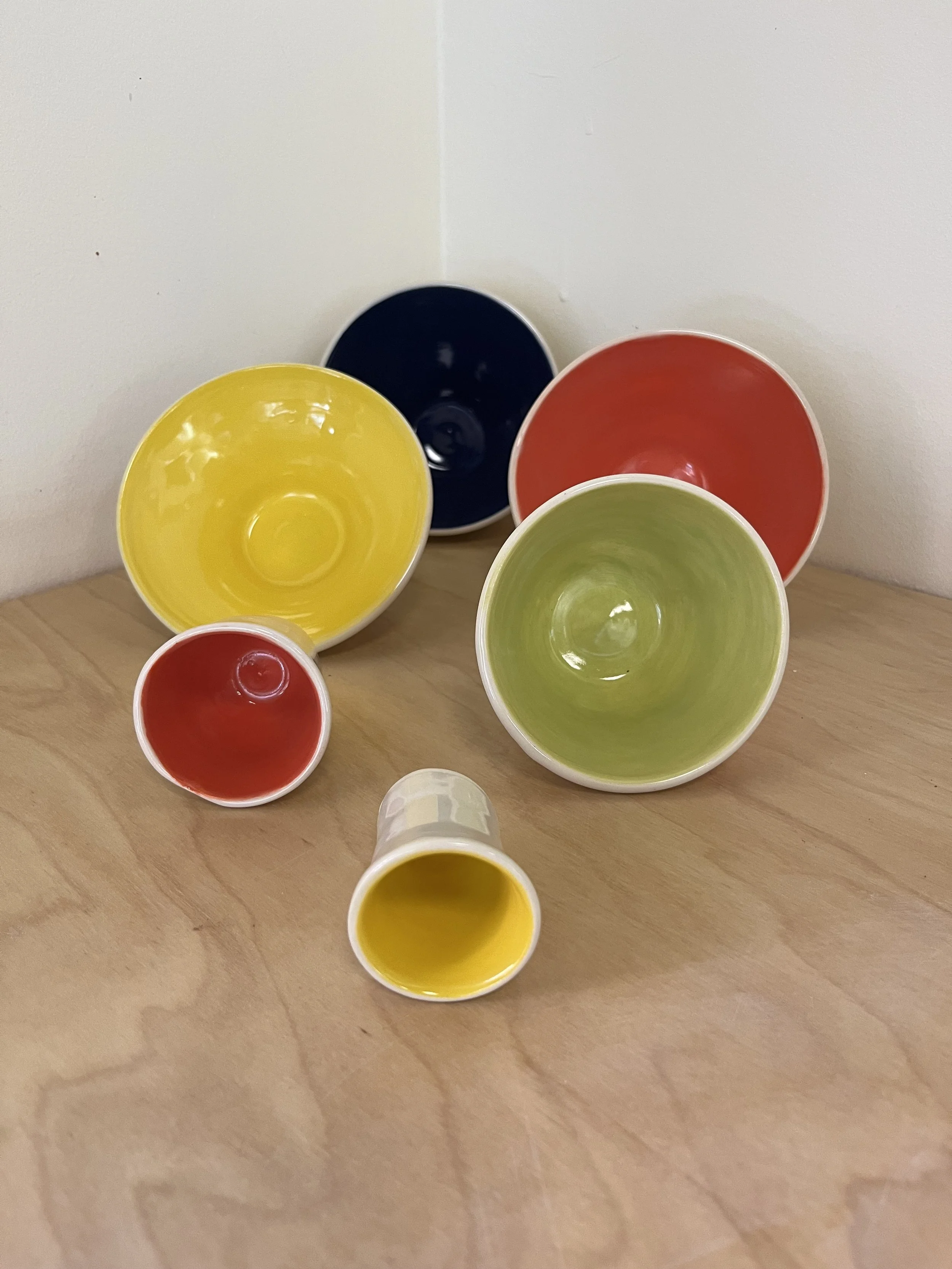 Six colorful bowls arranged on a wooden surface, with a plain white wall behind them. The bowls are red, yellow, green, and black, with some of each color visible.