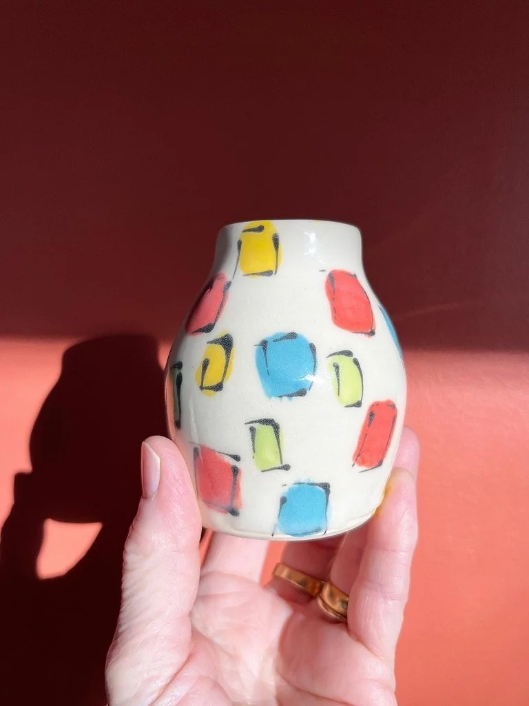 A small, white ceramic vase with colorful, abstract geometric patterns in red, blue, yellow, and black, held in a person's hand against a reddish background.