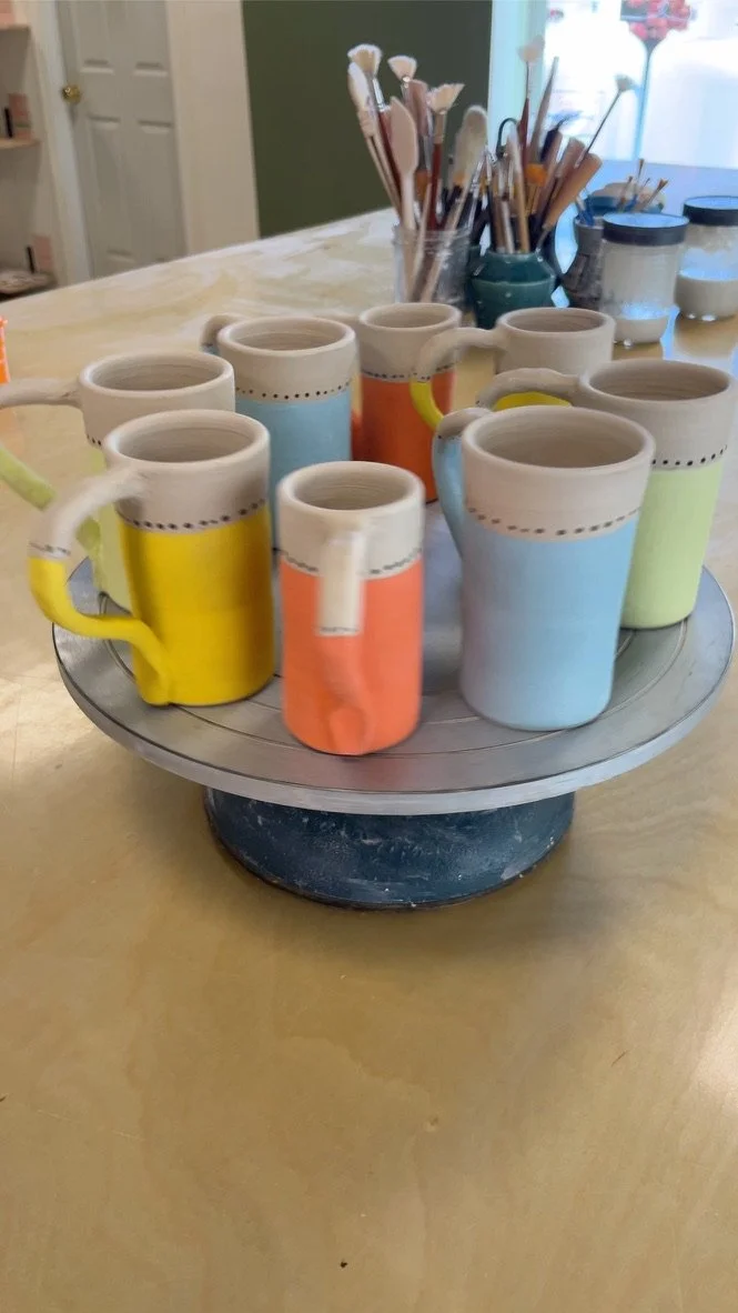 Colorful ceramic mugs on a round wooden display stand on a table, with paintbrushes and jars in the background.