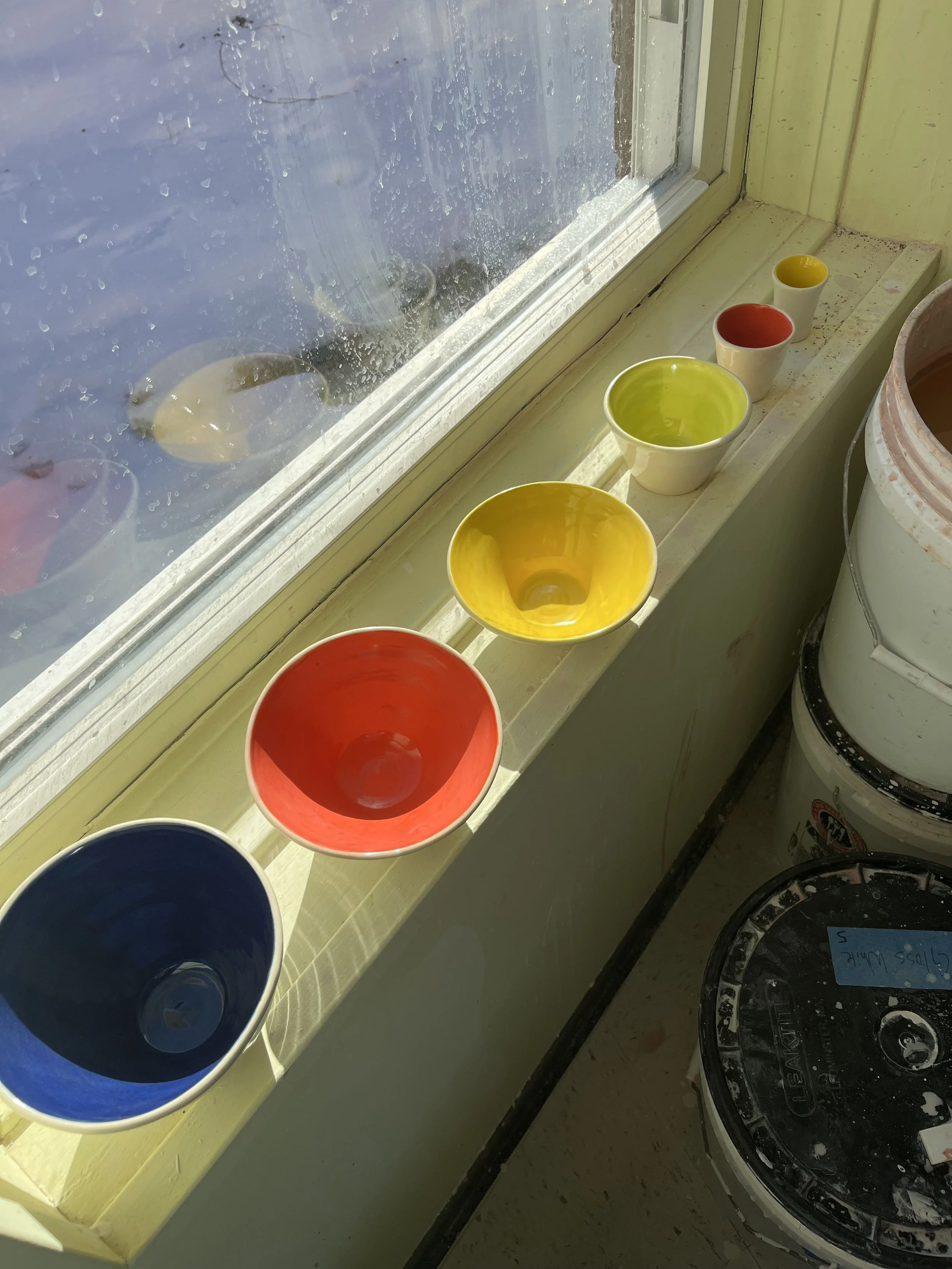 Colorful ceramic bowls in red, blue, yellow, green, and white on a windowsill, with a window showing raindrops outside and paint buckets below.