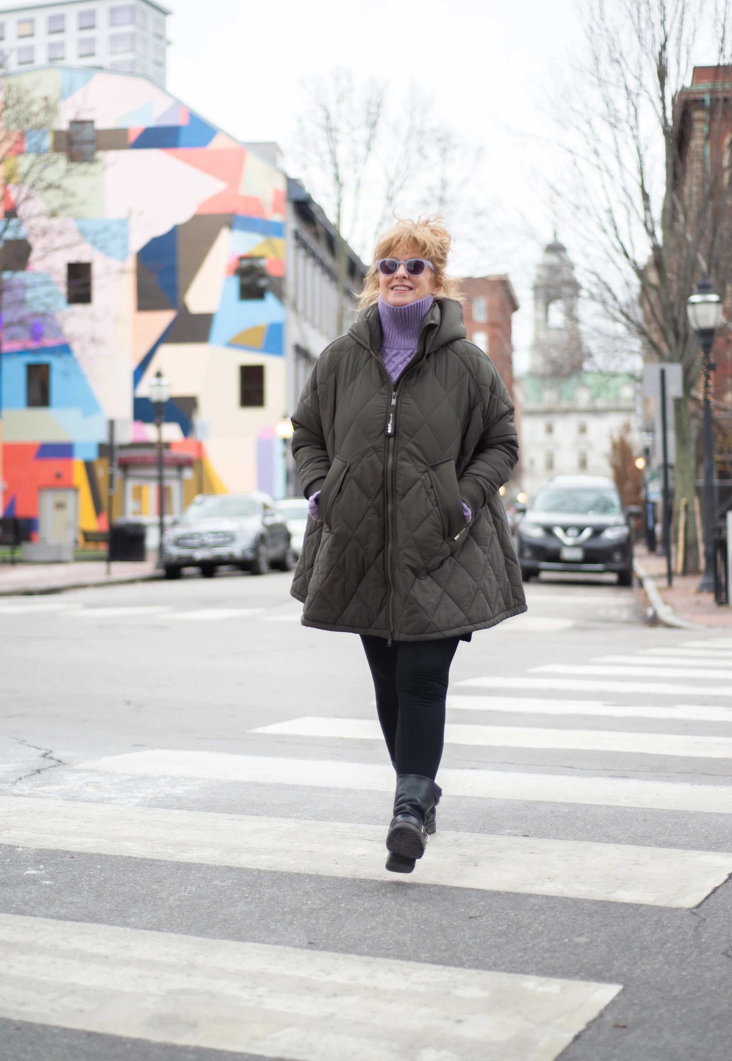 A woman walking across a crosswalk on a city street, wearing sunglasses, a purple turtleneck sweater, a dark green quilted coat, black leggings, and black boots, with a colorful geometric mural on the building in the background.