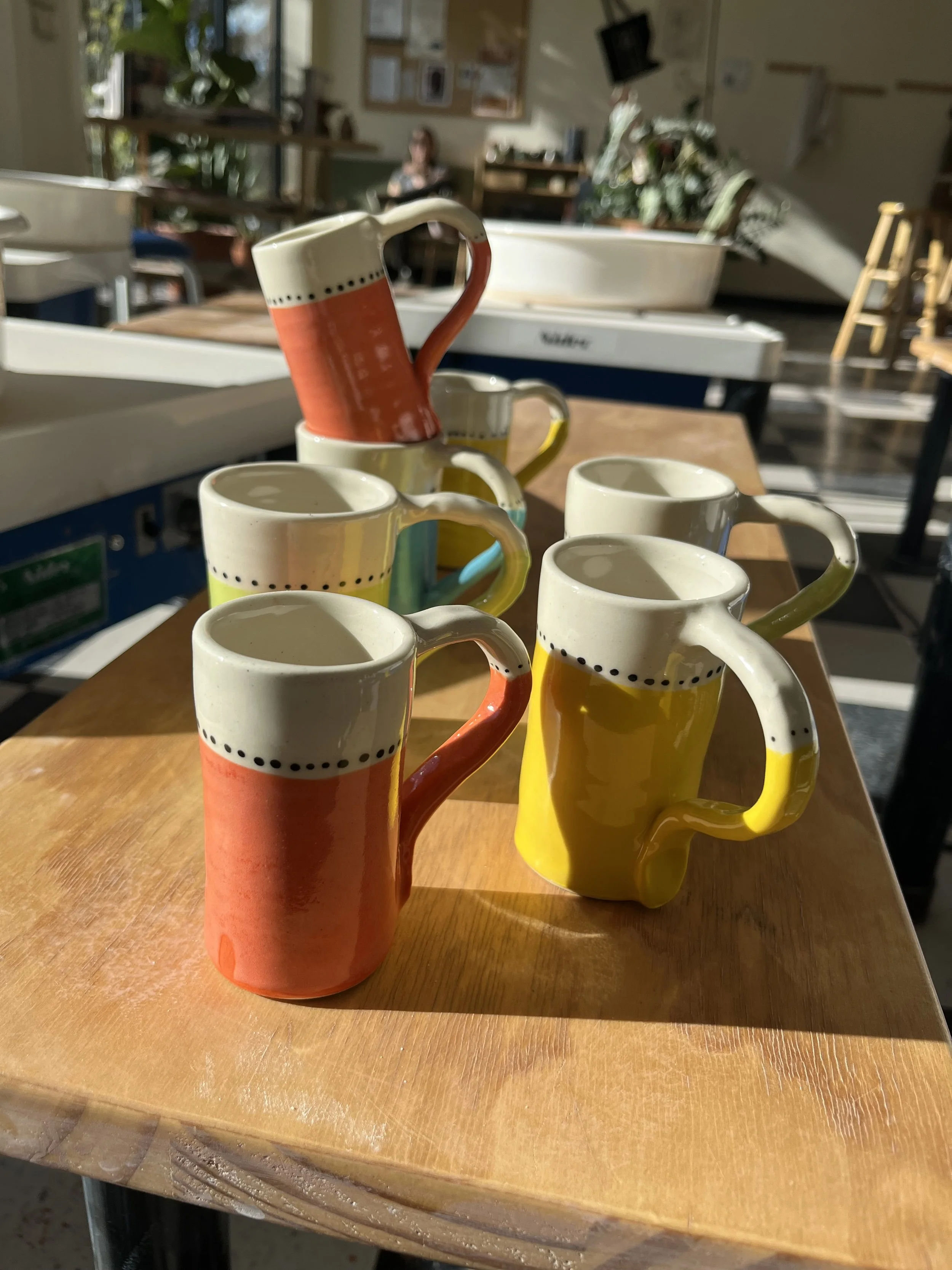 Six colorful ceramic mugs arranged on a wooden table with sunlight casting shadows, with a blurry background including a person sitting, plants, and various items in a room.