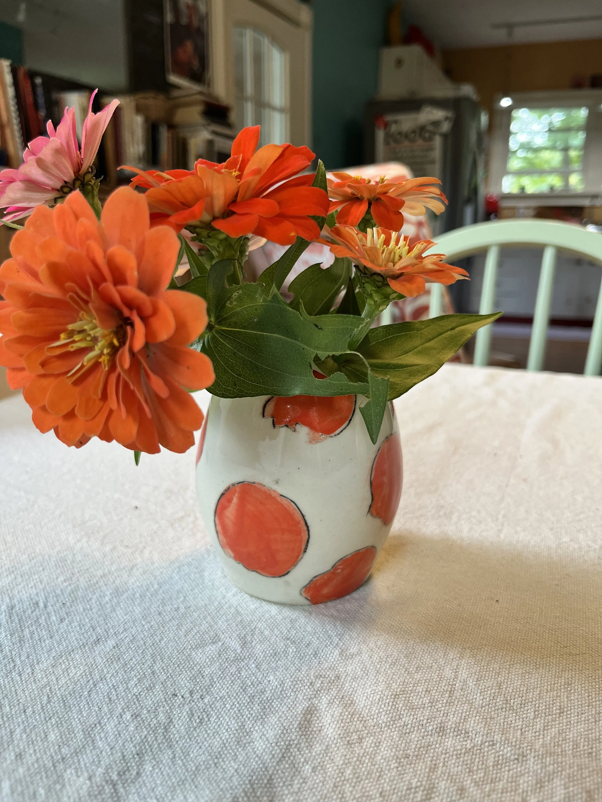 A small white vase with orange circles holds a bouquet of orange and pink flowers on a light-colored table inside a room with a window in the background.
