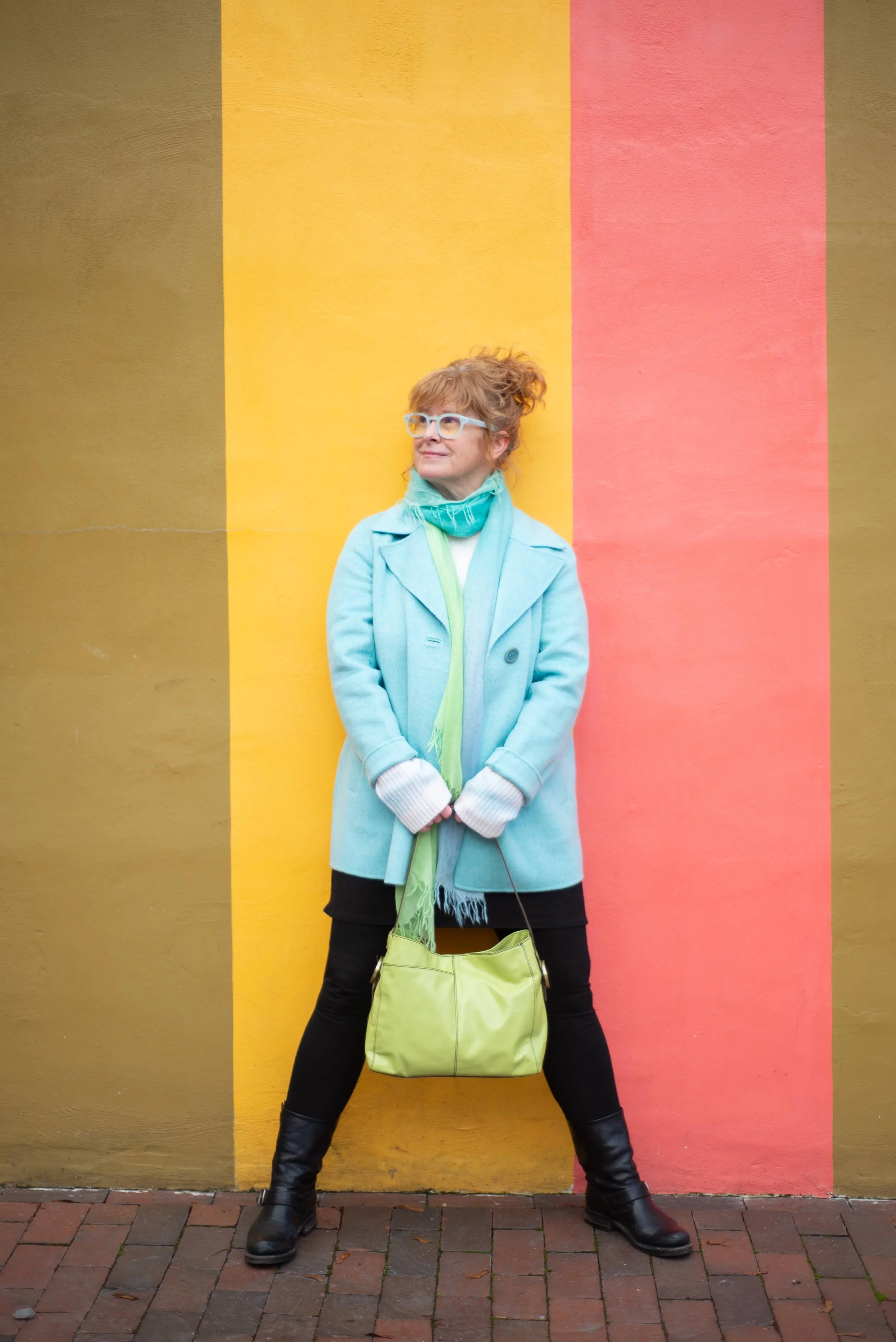 Woman standing against a wall painted with vertical stripes of pink, yellow, and brown, wearing a light blue coat, black pants, black boots, and holding a green bag.