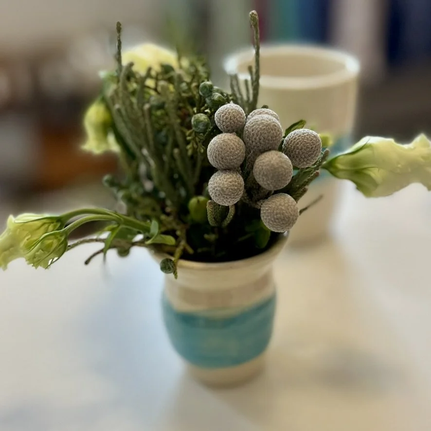 A small bouquet of artificial flowers in a white and blue ceramic vase, with a blurred background including a mug and some books.