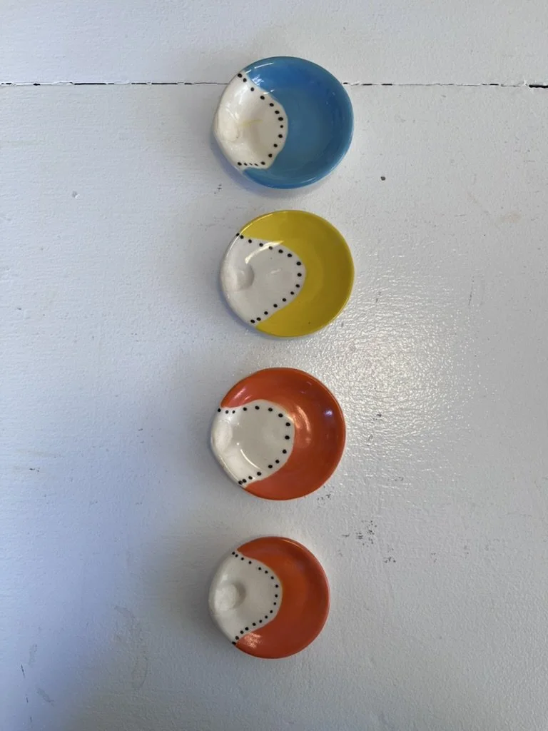 Four ceramic bowl-shaped objects in blue, yellow, and orange colors, arranged vertically. Each has a white, crescent-shaped section with black dotted details.