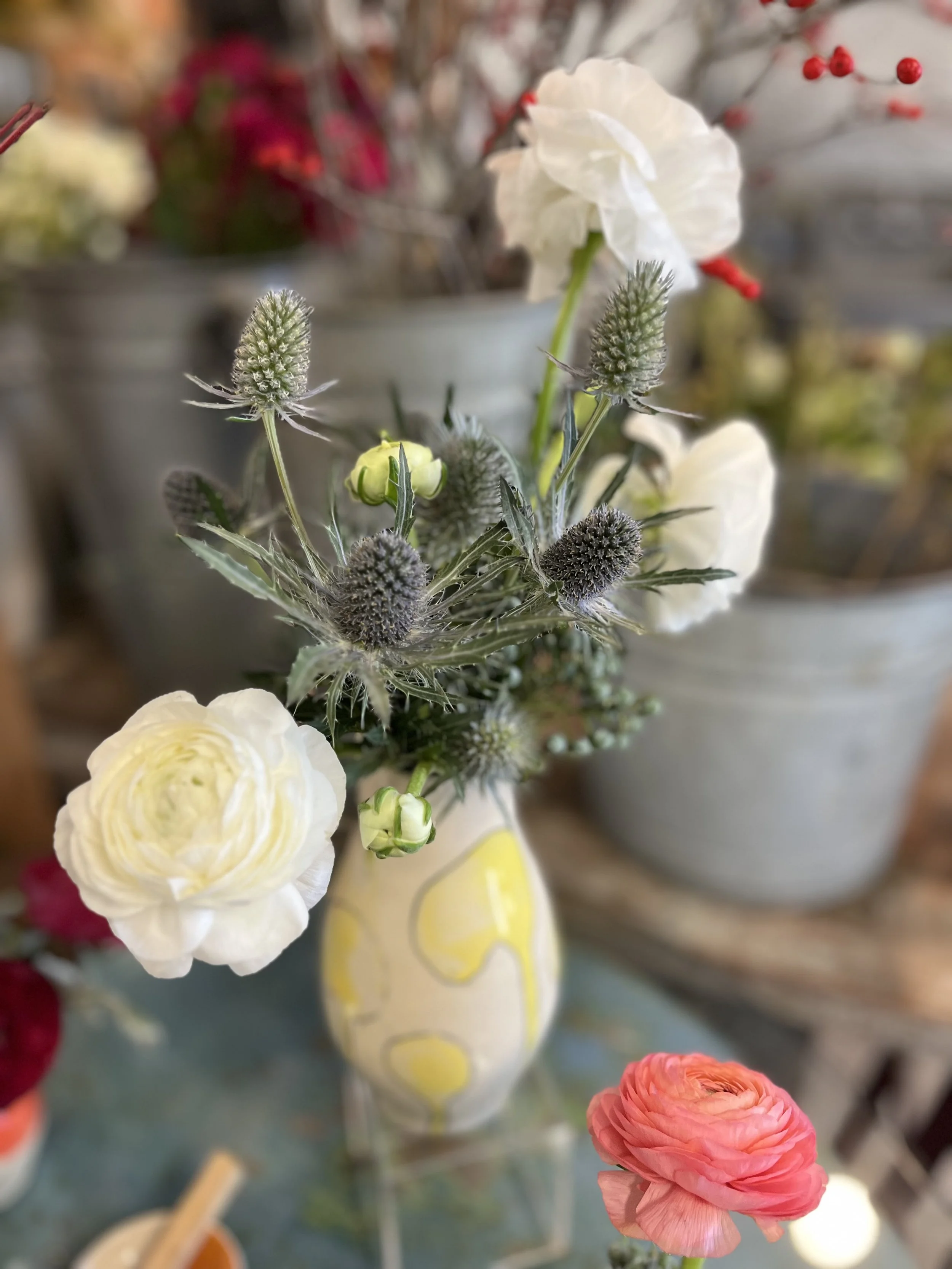 A close-up of a floral arrangement in a decorative vase with white, pink, and green flowers and gray botanical accents, with blurred background containers.